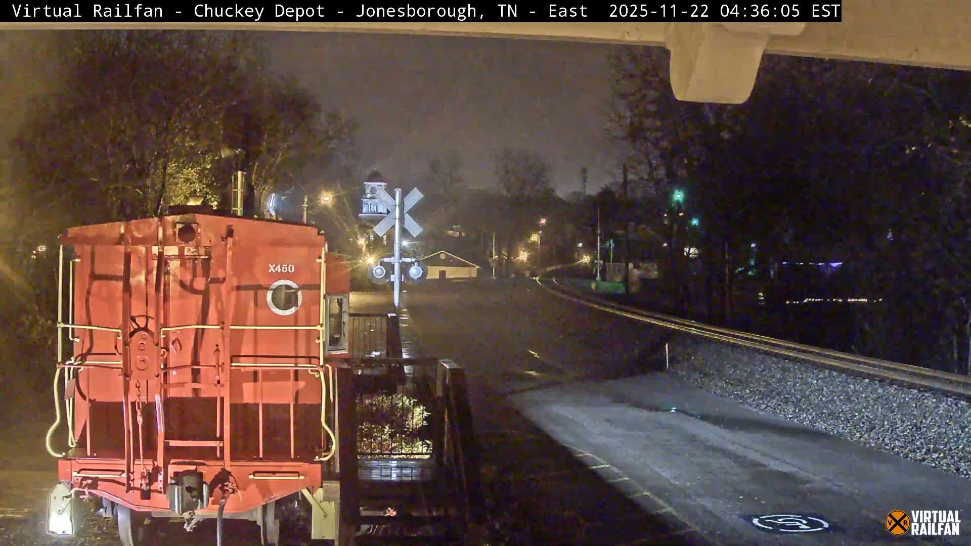 A red train engine marked "X450" sits at a railroad crossing on a wet, misty early morning, illuminated by streetlights with tracks diverging into the distance and buildings faintly visible through the haze.