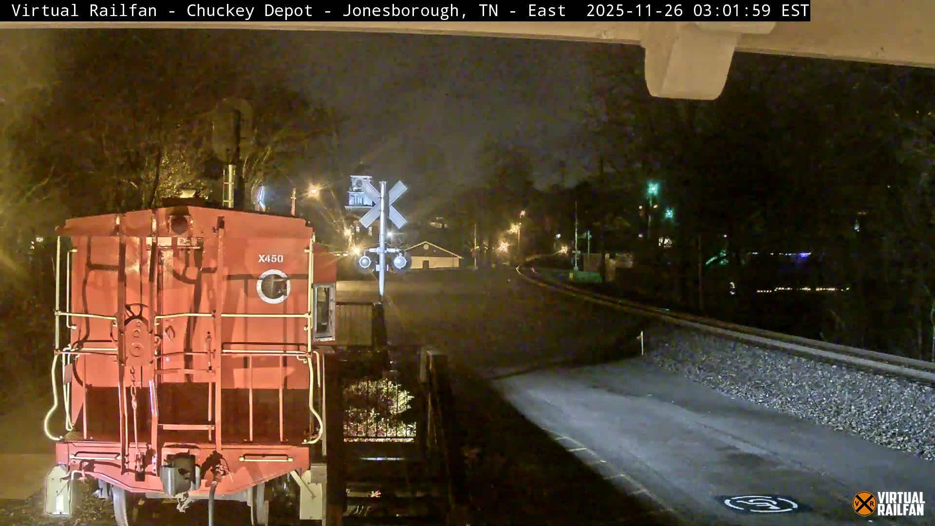 A red train is parked next to a railroad crossing sign at night, with tracks curving into the distance under an overcast sky illuminated by scattered lights.