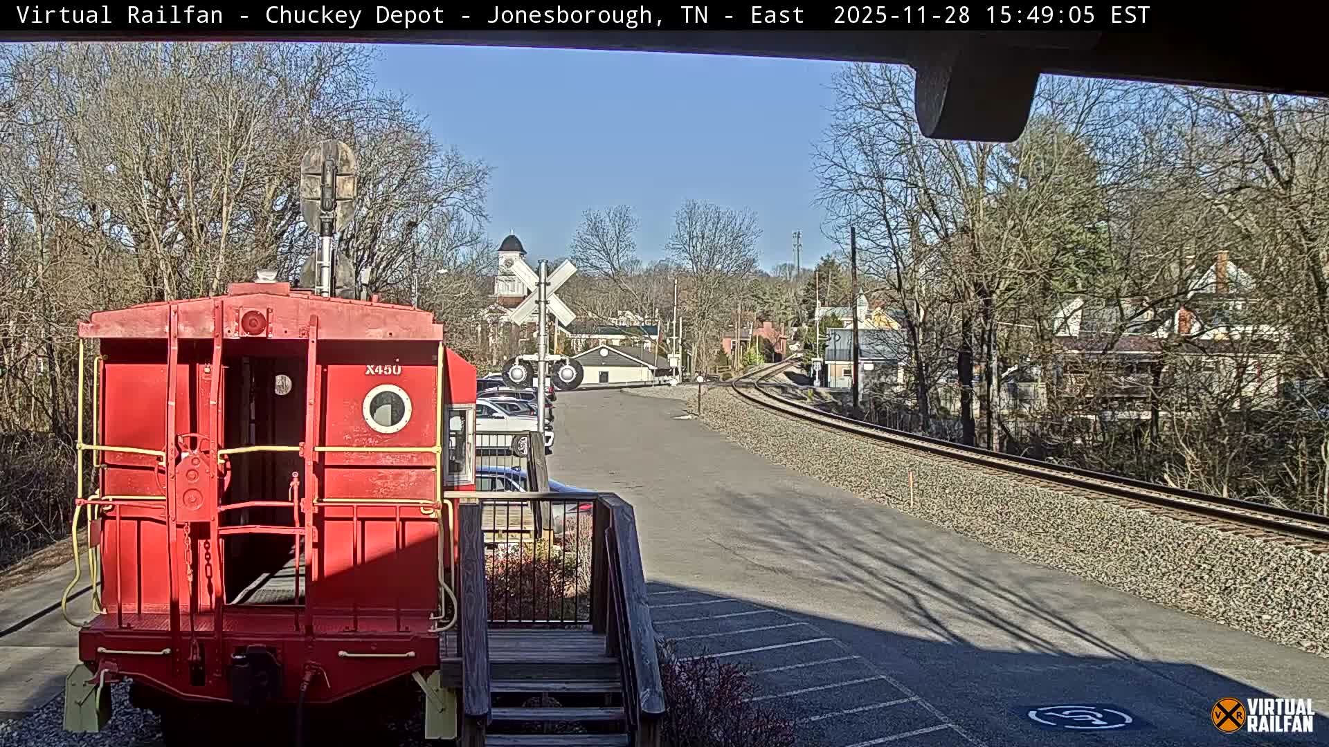A bright red caboose stands prominently in the foreground beside a wooden deck and train tracks that curve into the distance, with a clear blue sky overhead and bare trees framing a small town in the background under sunny conditions.