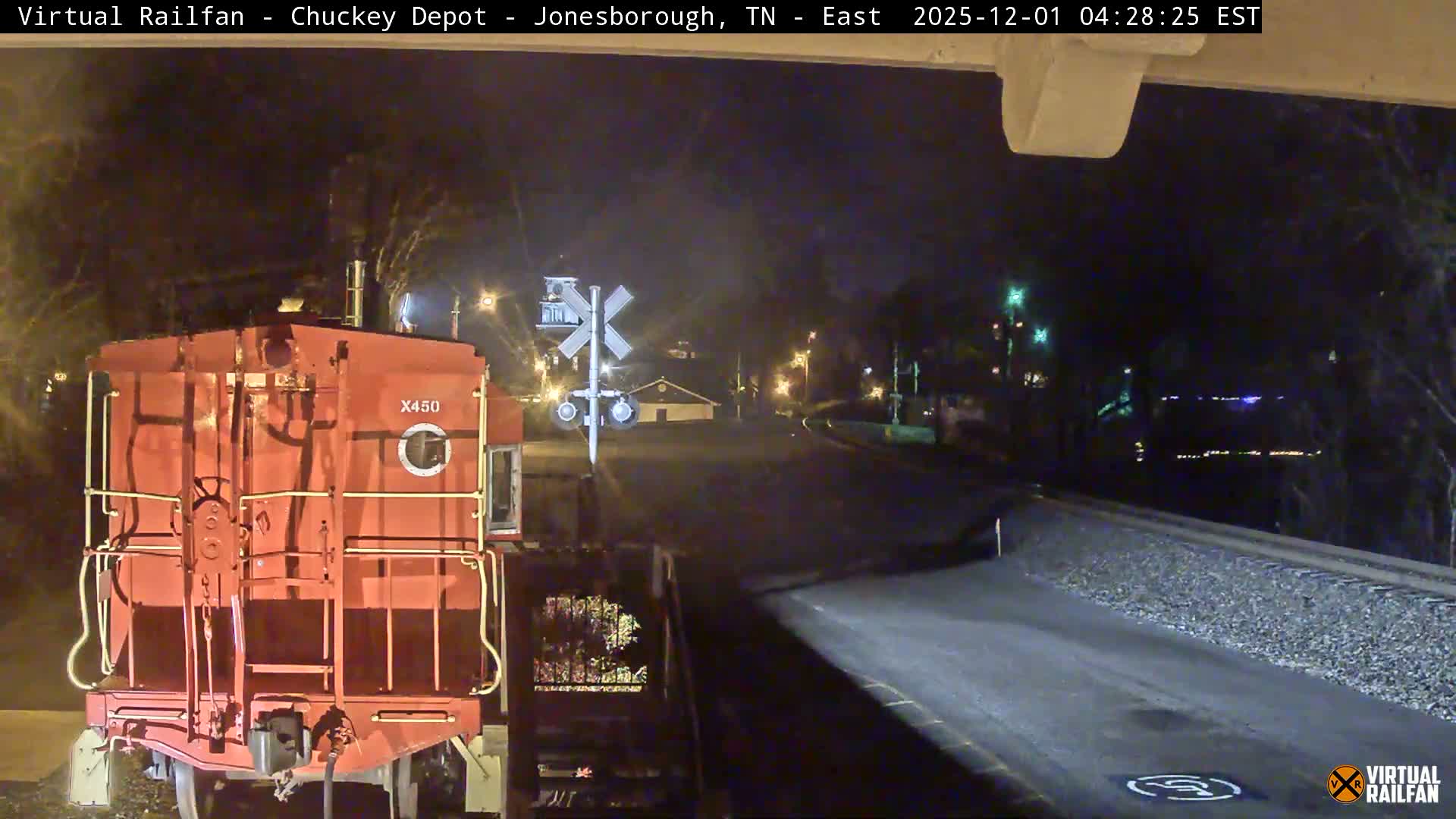 A bright red train car stands next to a railroad crossing with flashing lights, alongside a curving, streetlight-lit road and railroad tracks on a clear night.
