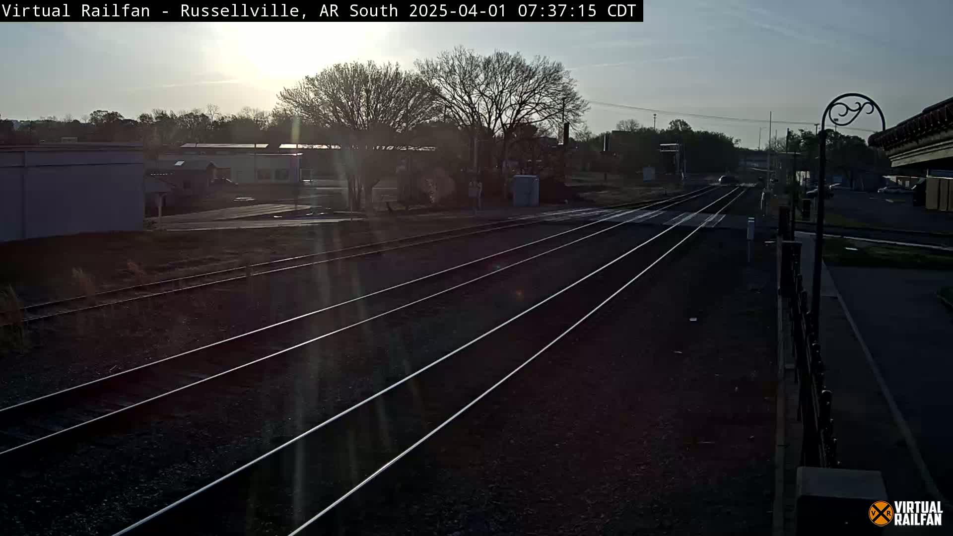 A view of train tracks on a sunny morning, with buildings and trees visible in the background.