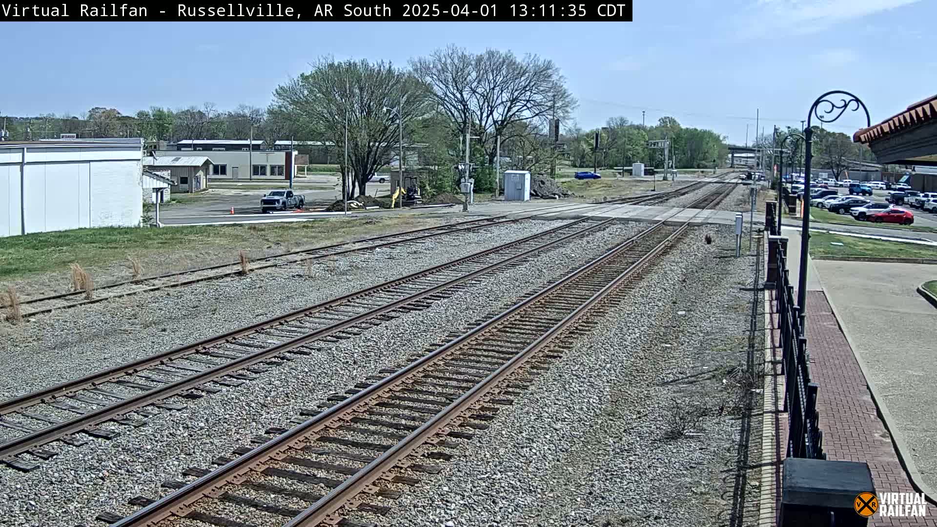 A sunny day reveals multiple train tracks extending into the distance, passing by buildings and parked cars.
