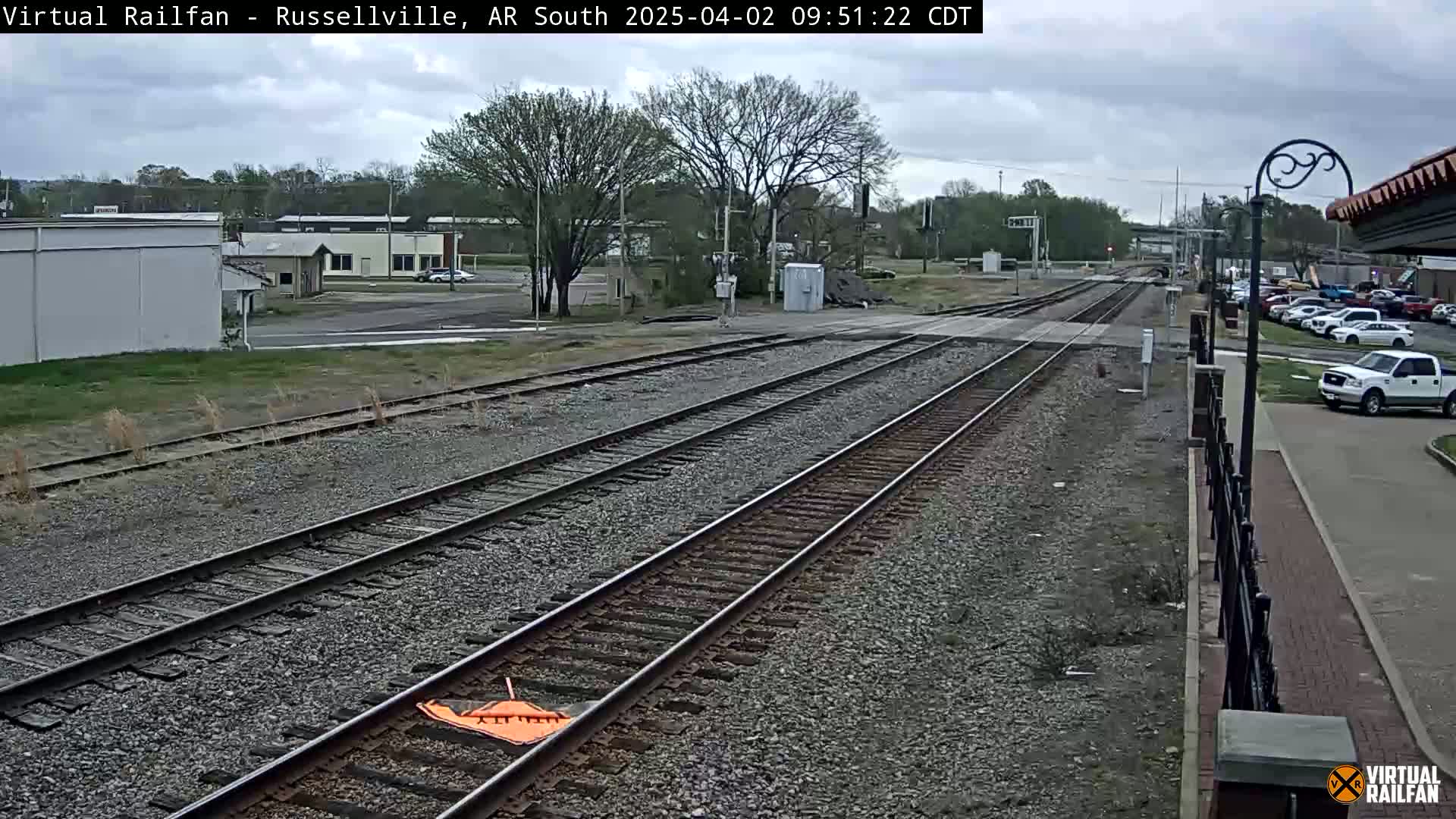 Multiple train tracks stretch into the distance under an overcast sky, past a small town with buildings and parked cars.