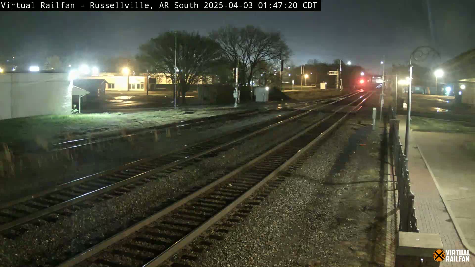 A nighttime view of a train passing through a railway station, with wet ground indicating recent rain.