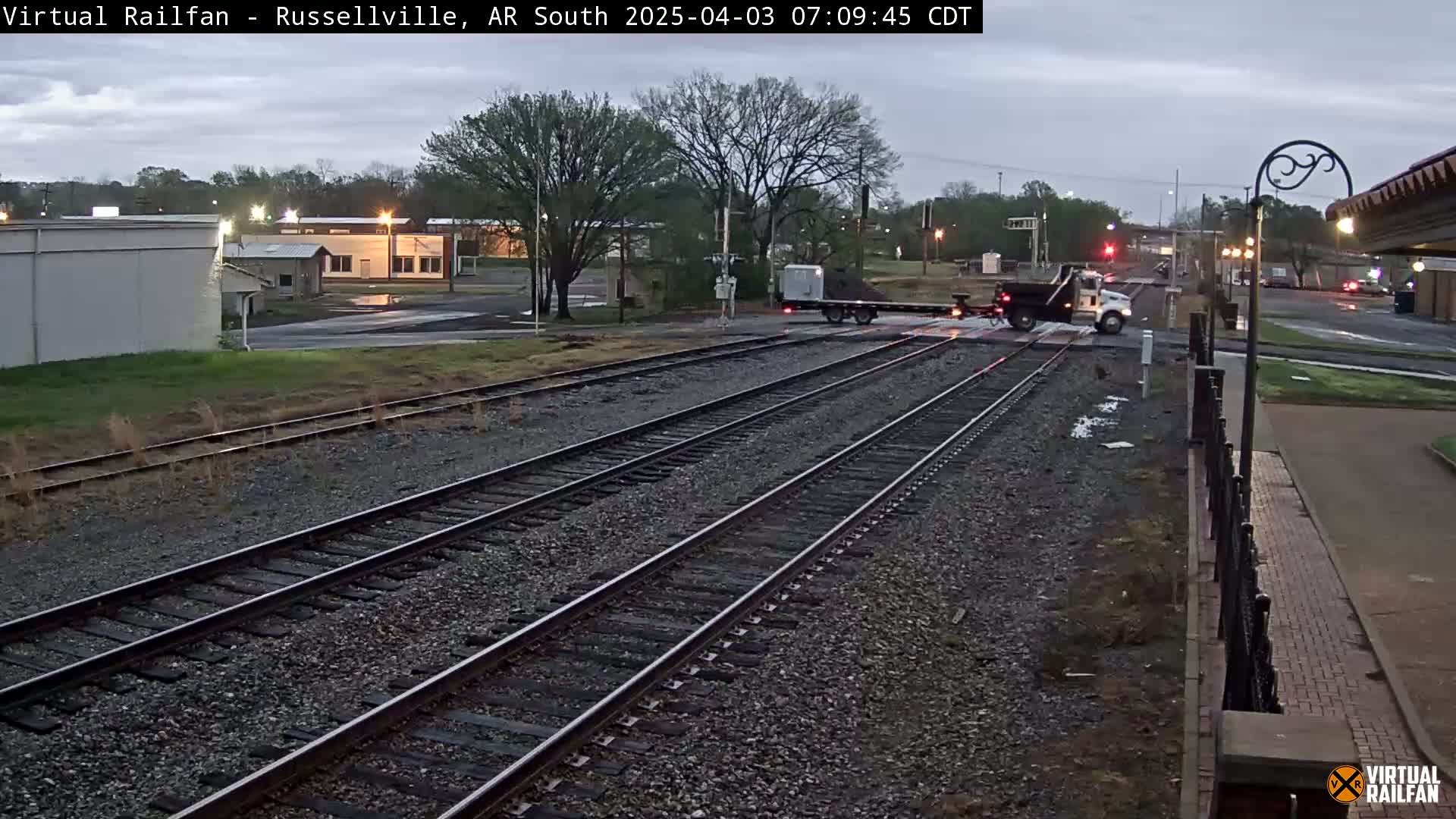 A dump truck and a flatbed truck are crossing railroad tracks in an overcast urban setting.