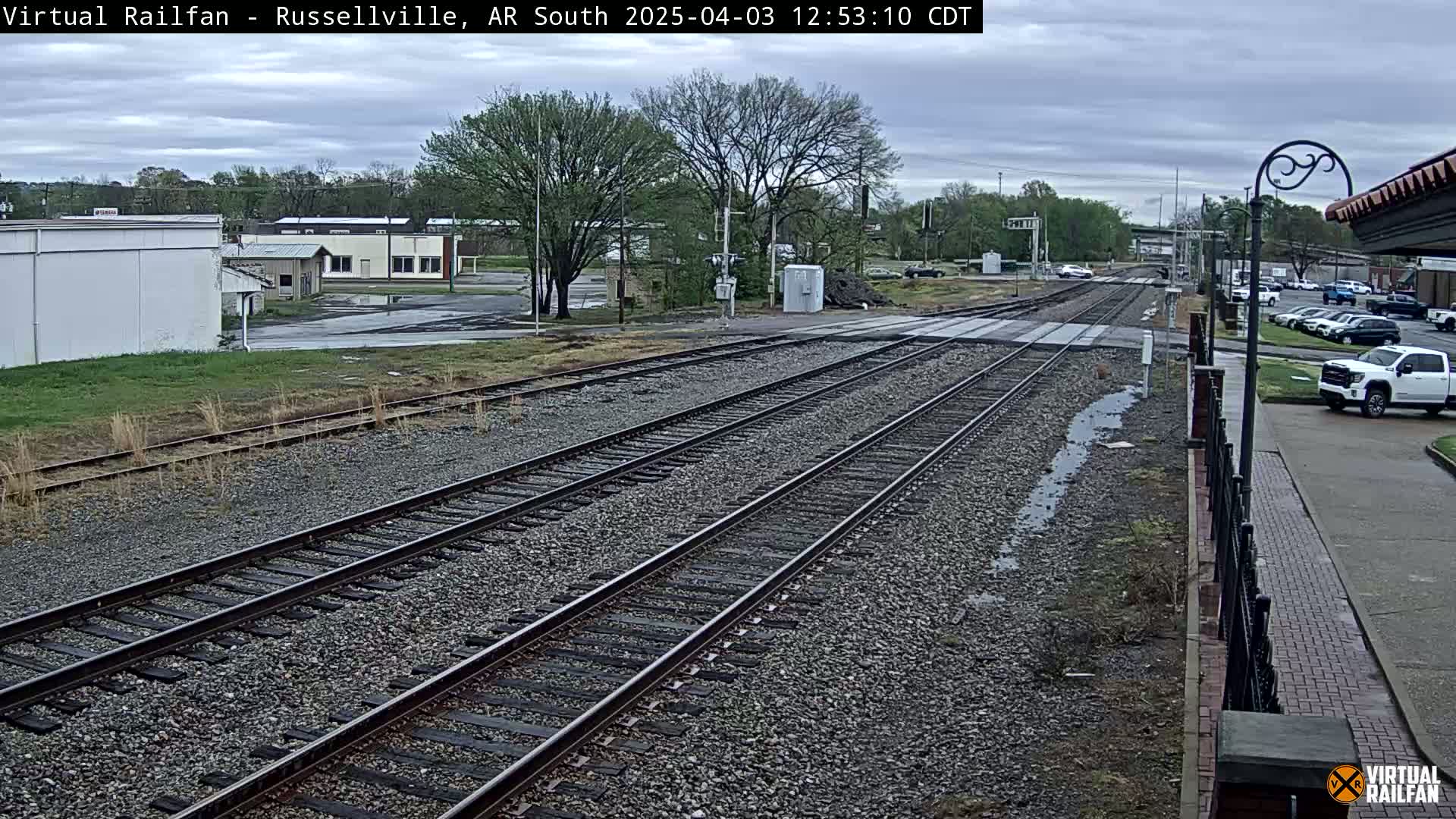 Multiple train tracks stretch into the distance under an overcast sky, passing near a few buildings and a parking lot with several cars.