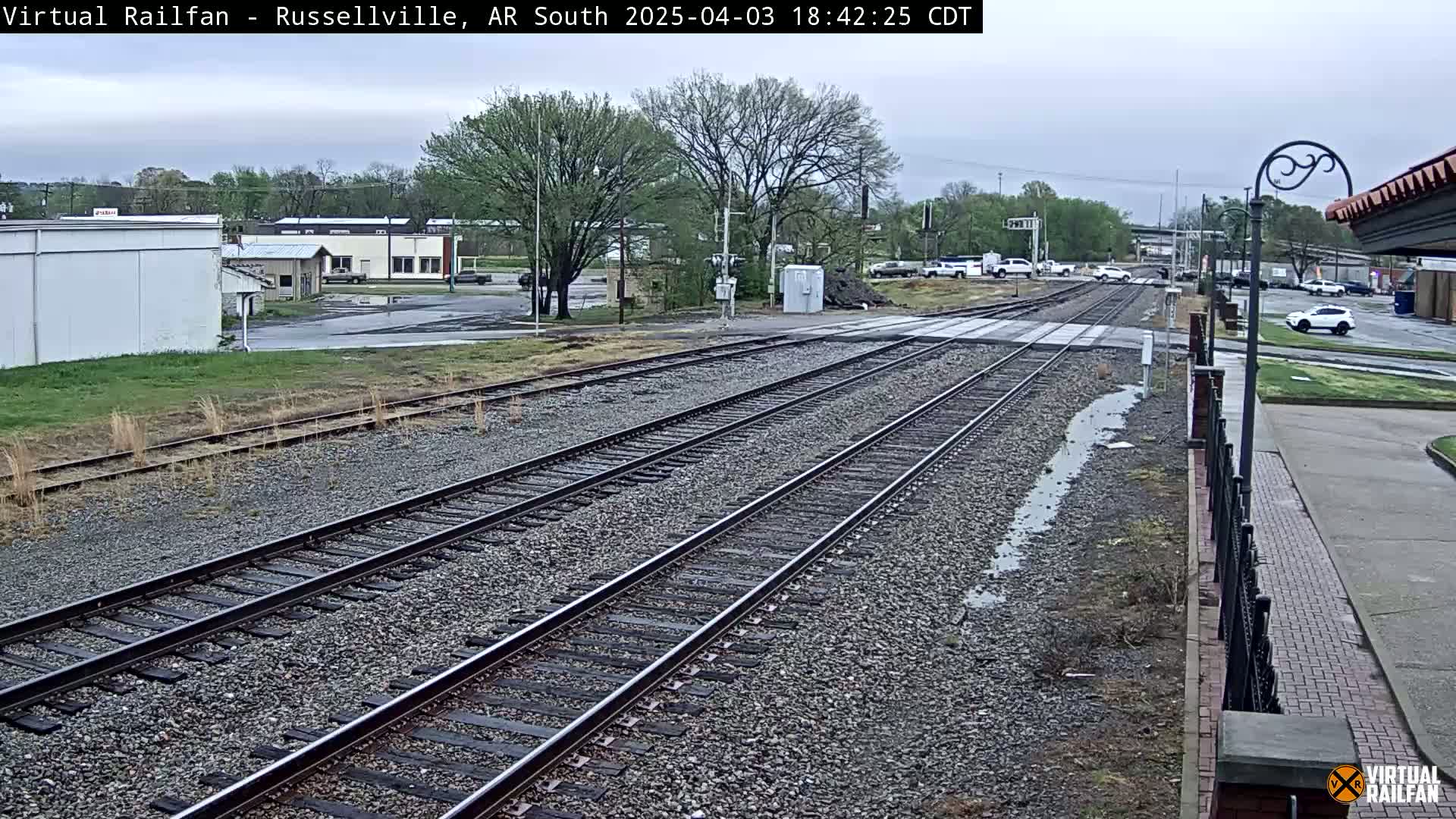 Multiple train tracks extend into the distance, past a street with cars parked along the side, under an overcast sky.