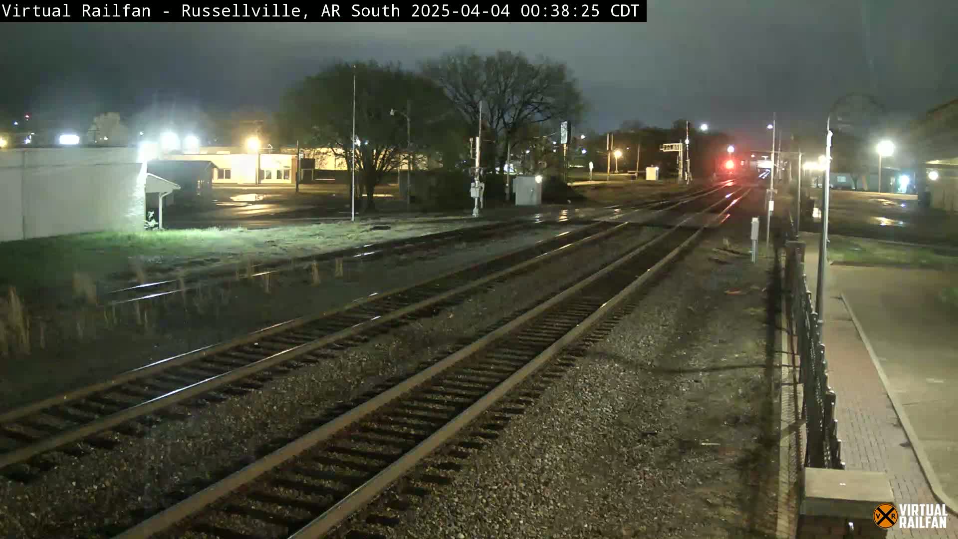 A nighttime view of train tracks, wet from recent rain, with a train in the distance and buildings and trees visible in the background.