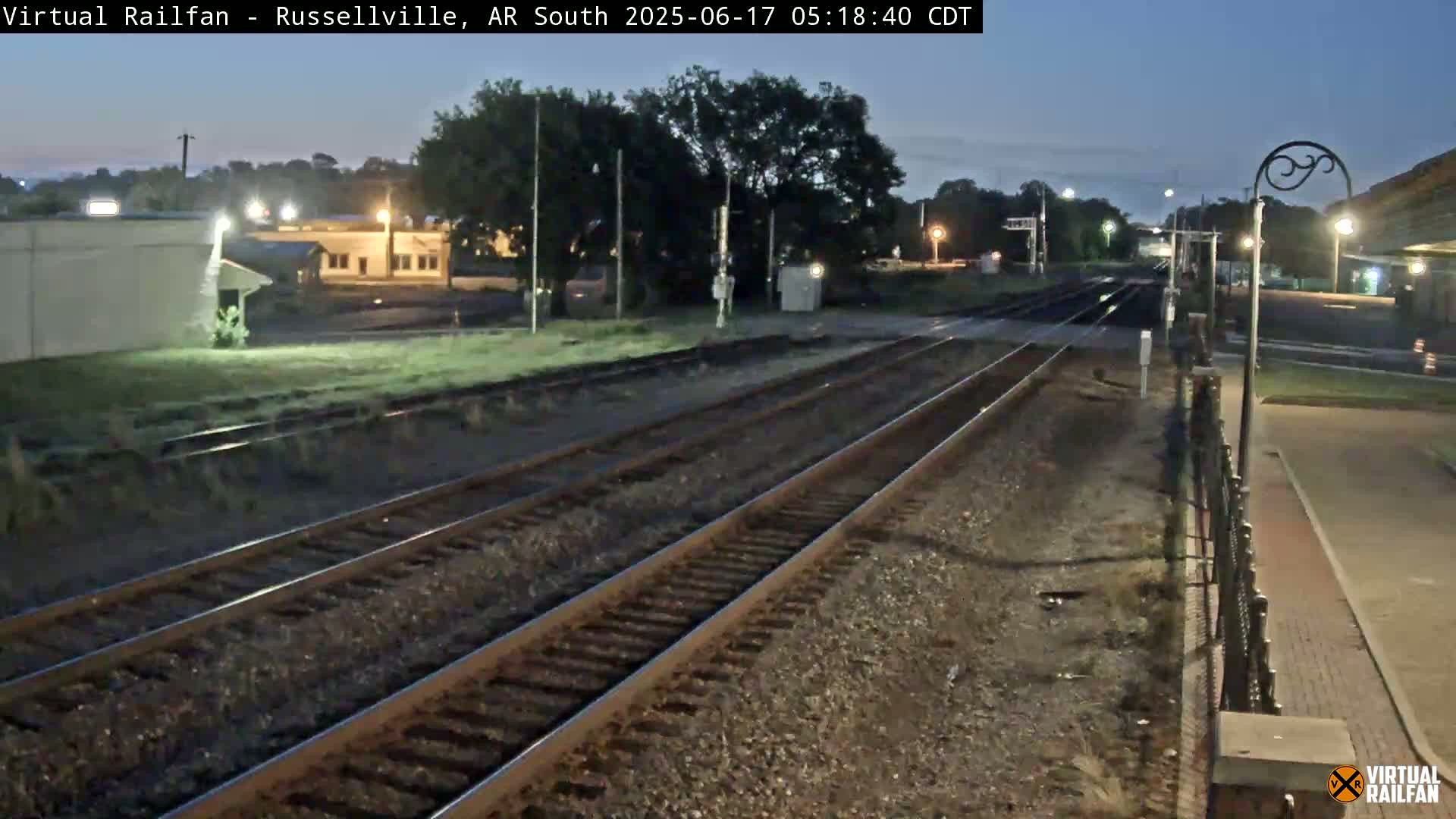 Under a dark, twilight sky, two sets of train tracks extend into the distance, passing by a small town with dimly lit buildings.