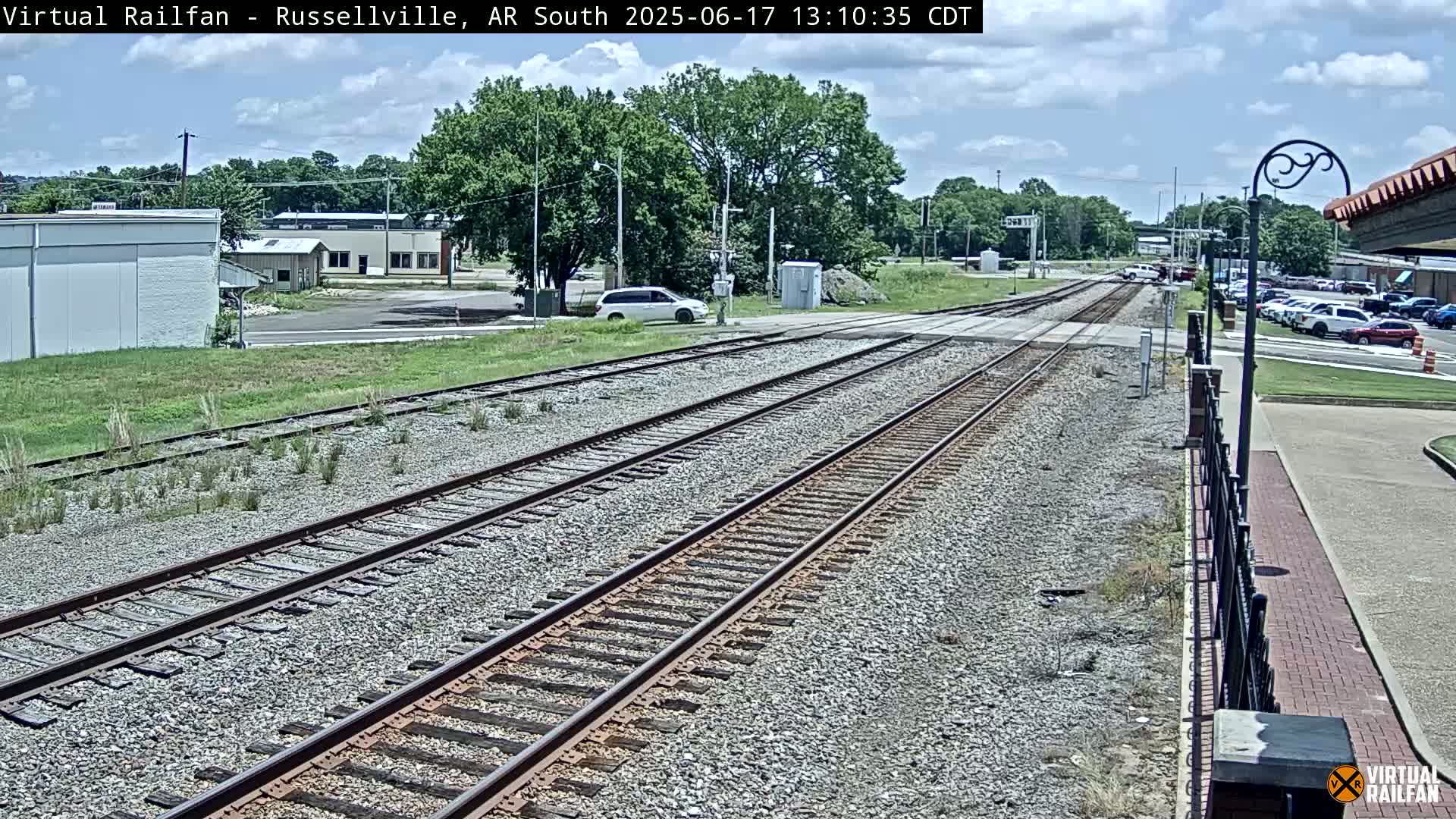 A mostly sunny day reveals a view of railroad tracks, a nearby road with cars parked and a few buildings in the distance.