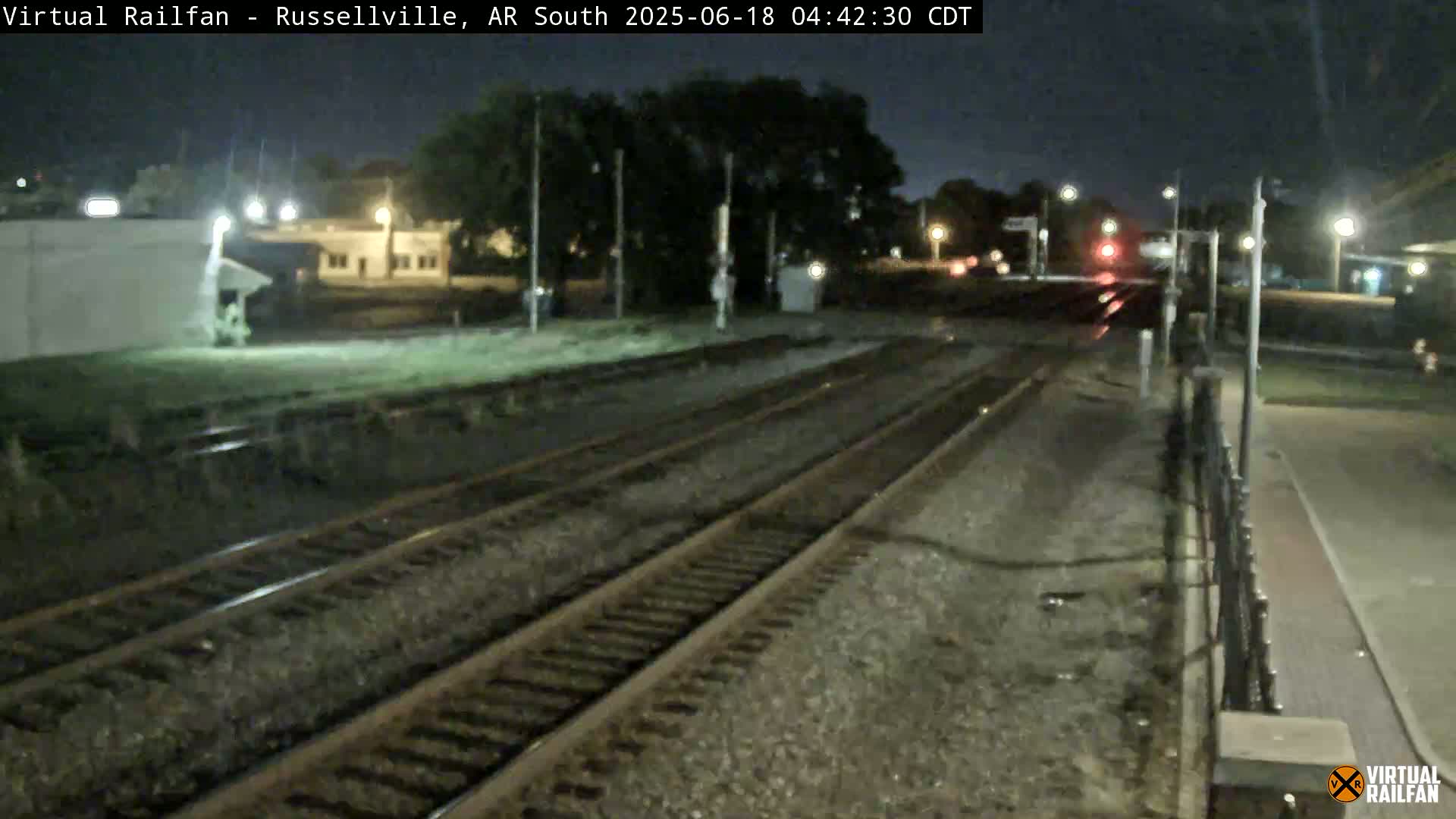 A nighttime view of railroad tracks at a station, with buildings and trees visible in the background under a dark sky.