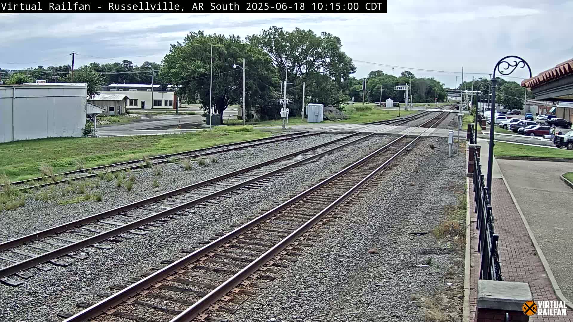 Multiple train tracks extend into the distance on a partly cloudy day, with a few buildings and parked cars visible in the background.