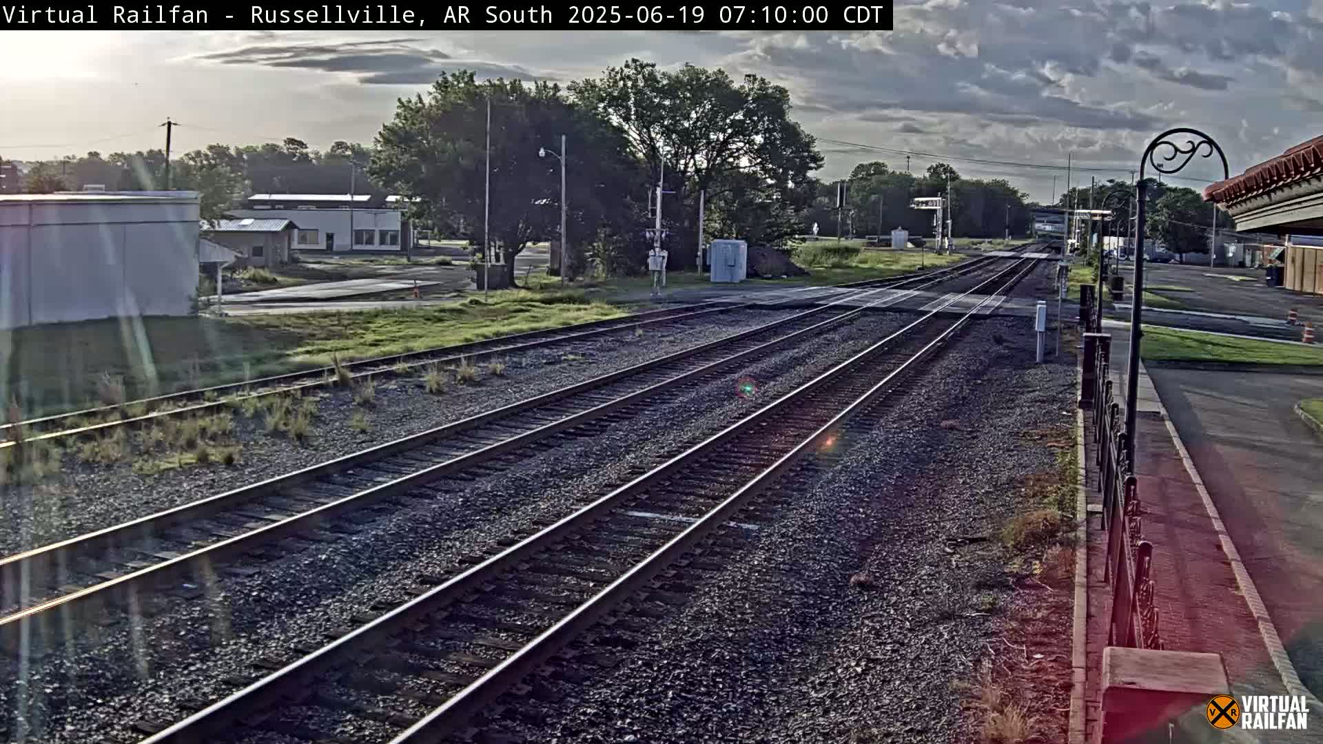 Multiple train tracks stretch into the distance under a partly cloudy sky, passing by some low buildings and green vegetation.