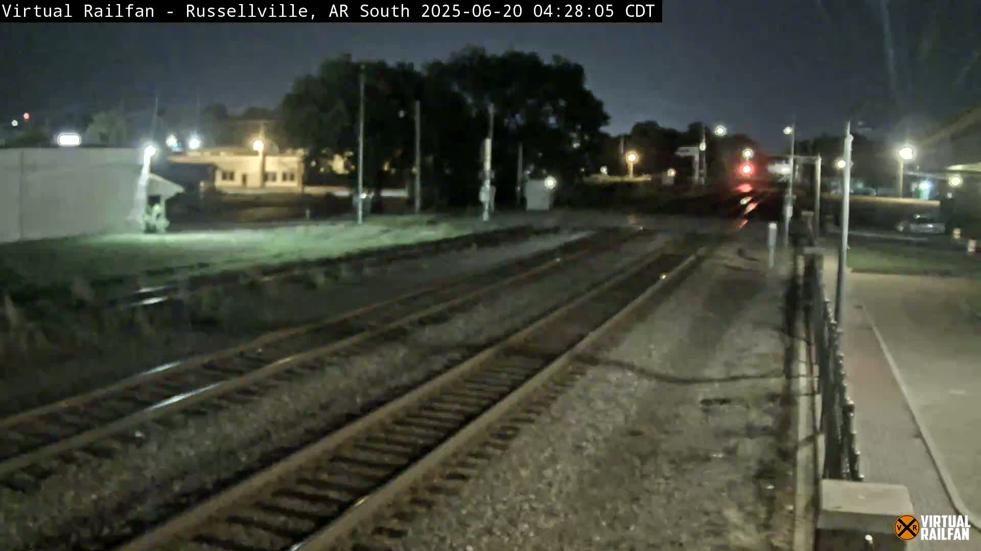 A nighttime view of train tracks at a station, with a train passing in the distance under dark, clear skies.