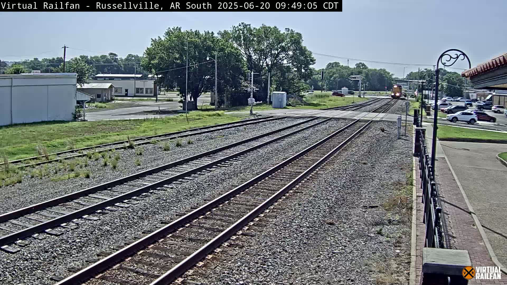 A train approaches a railway crossing on a sunny day, with buildings and parked cars visible in the background.