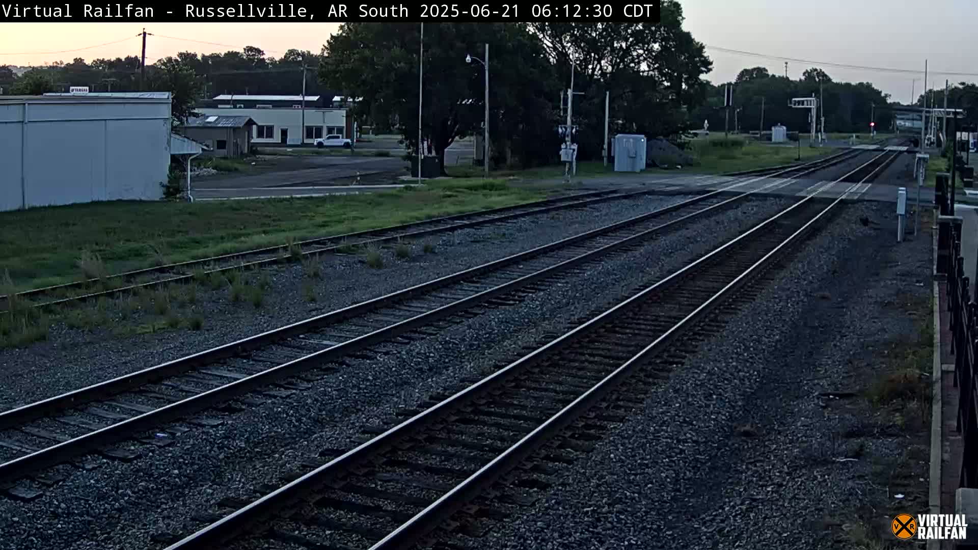 Three sets of train tracks extend into the distance under a twilight sky, passing by buildings and some sparse vegetation.