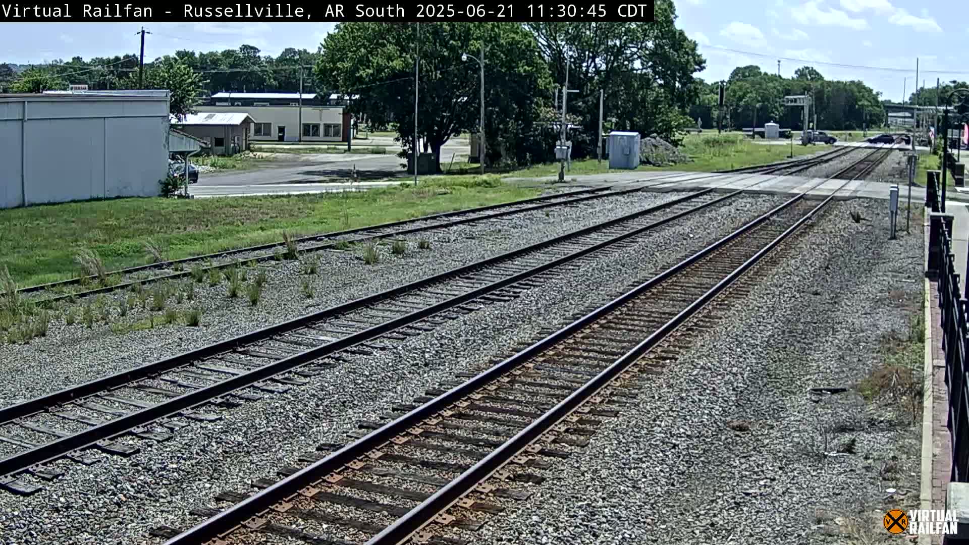 Multiple train tracks stretch from a road crossing into the distance under a partly sunny sky, with buildings and trees visible in the background.