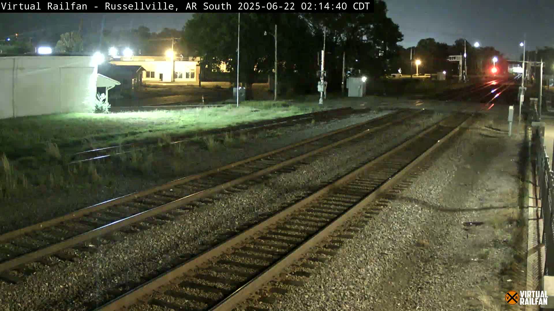 A nighttime view of train tracks, with some buildings and vegetation visible in the background under dark conditions.