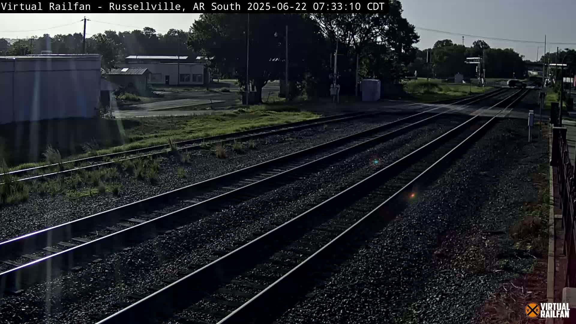 Multiple train tracks extend into the distance on a sunny day, past a roadside with buildings and trees.