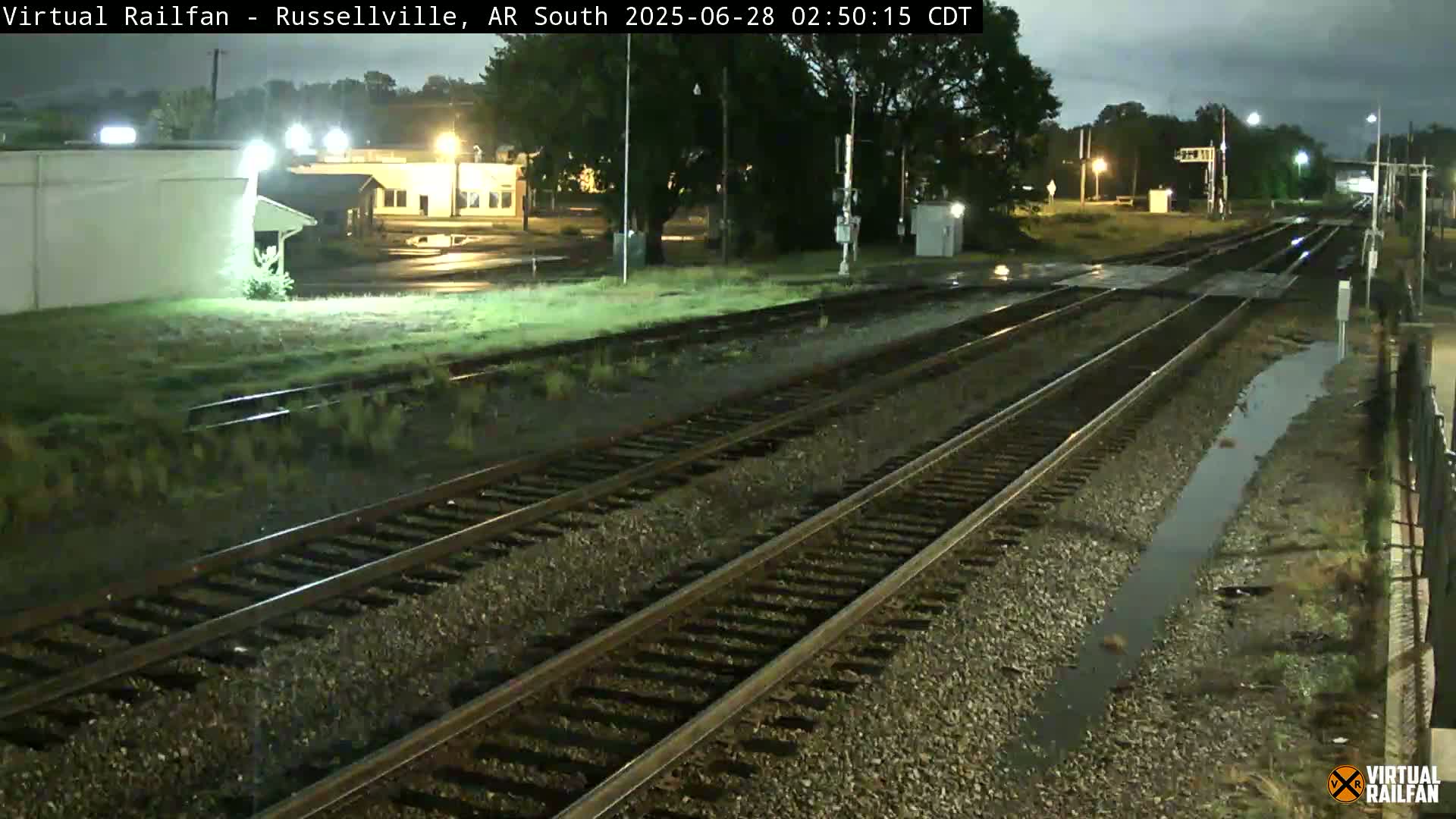 A nighttime view of railroad tracks in the rain, with some buildings and vegetation visible in the background.