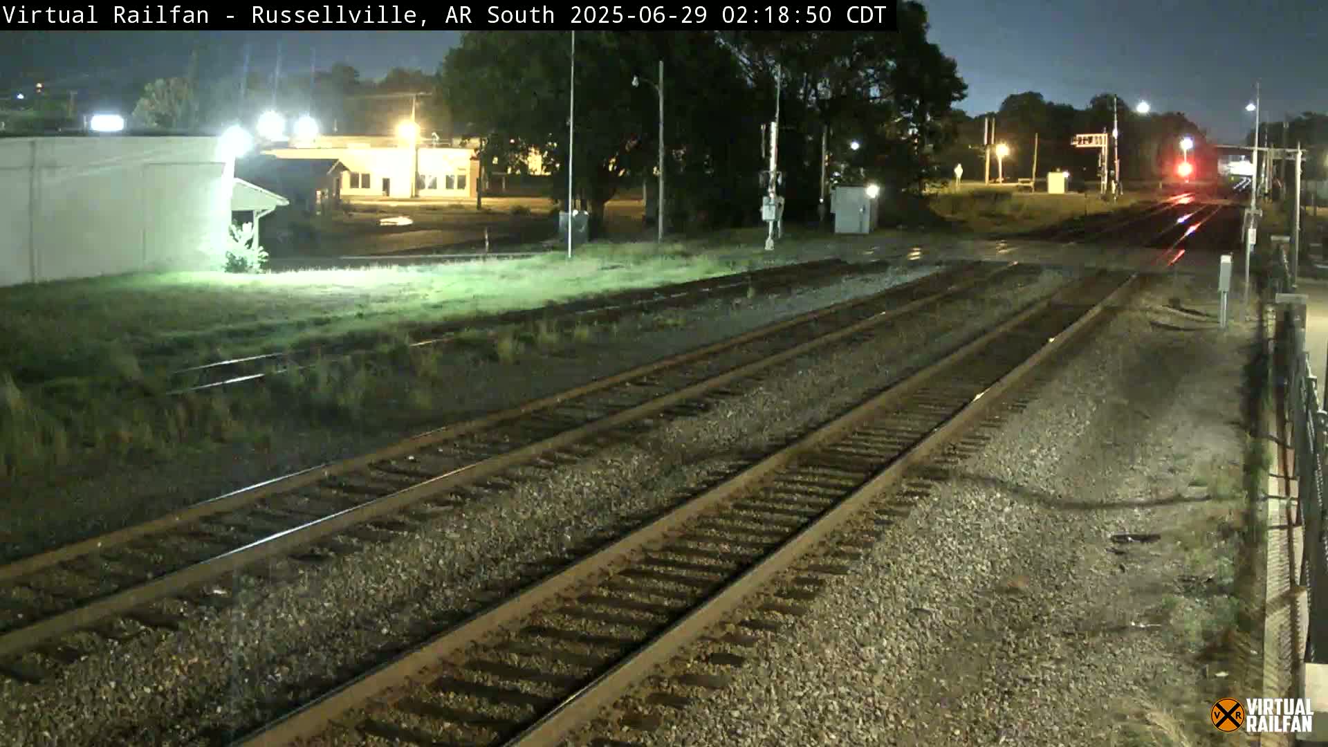 A nighttime view of railroad tracks extending into the distance, passing near some buildings under partly cloudy skies.