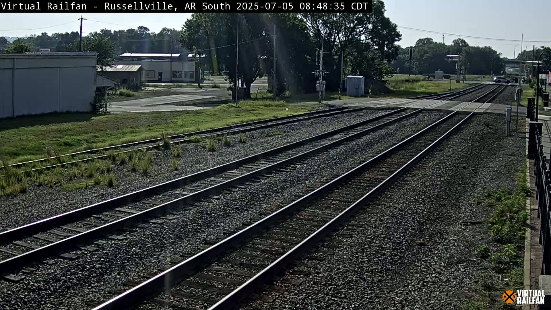 Multiple train tracks stretch into the distance on a sunny day, passing through a sparsely populated area with low buildings and green vegetation.