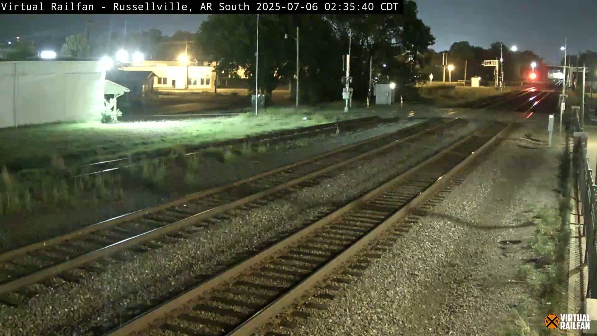 A nighttime view of railroad tracks at a station, with some buildings visible in the background under a dark sky.