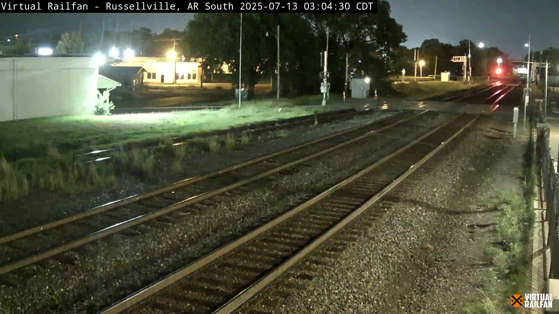 A nighttime view of railroad tracks, wet from recent rain, stretches towards a distant, faintly lit area with buildings and trees.