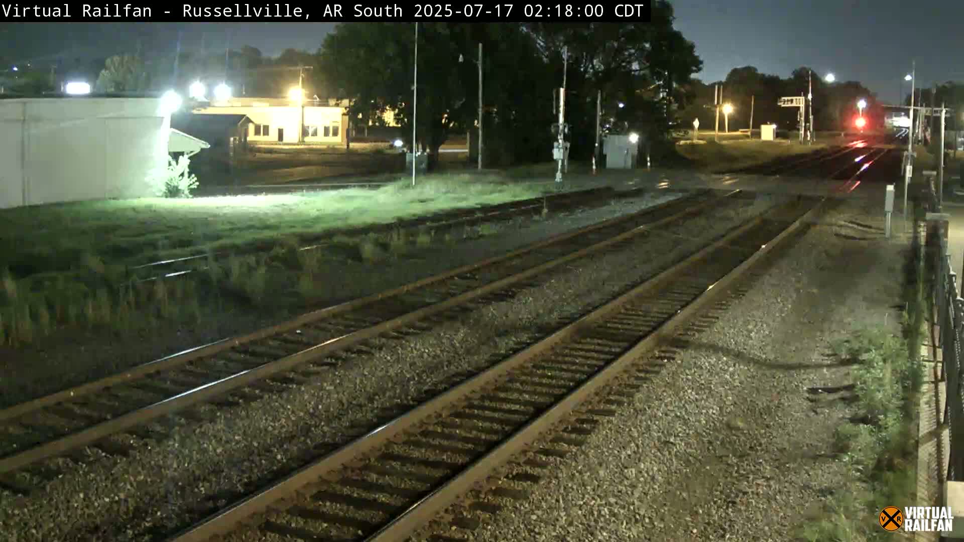 At night, multiple train tracks extend into the distance, alongside a grassy area and some buildings under a dark sky.