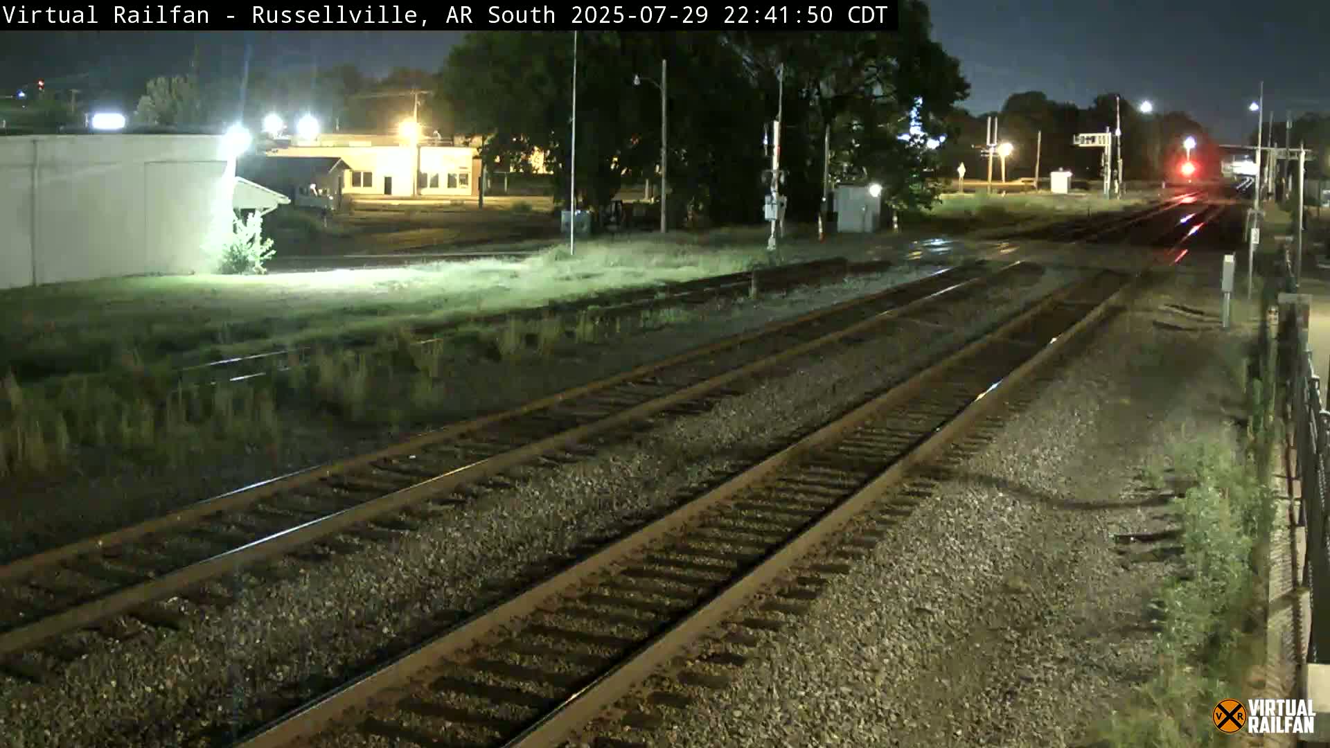 At night, multiple train tracks stretch into the distance past a grassy area and buildings under a clear, dark sky.