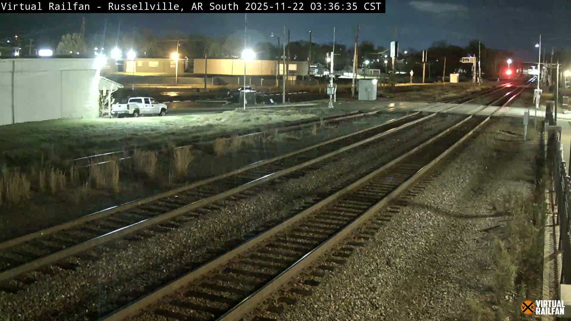 Multiple railroad tracks extend through an illuminated outdoor night scene, featuring streetlights, buildings, a white pickup truck, and a prominent red signal light in the distance under a clear sky.
