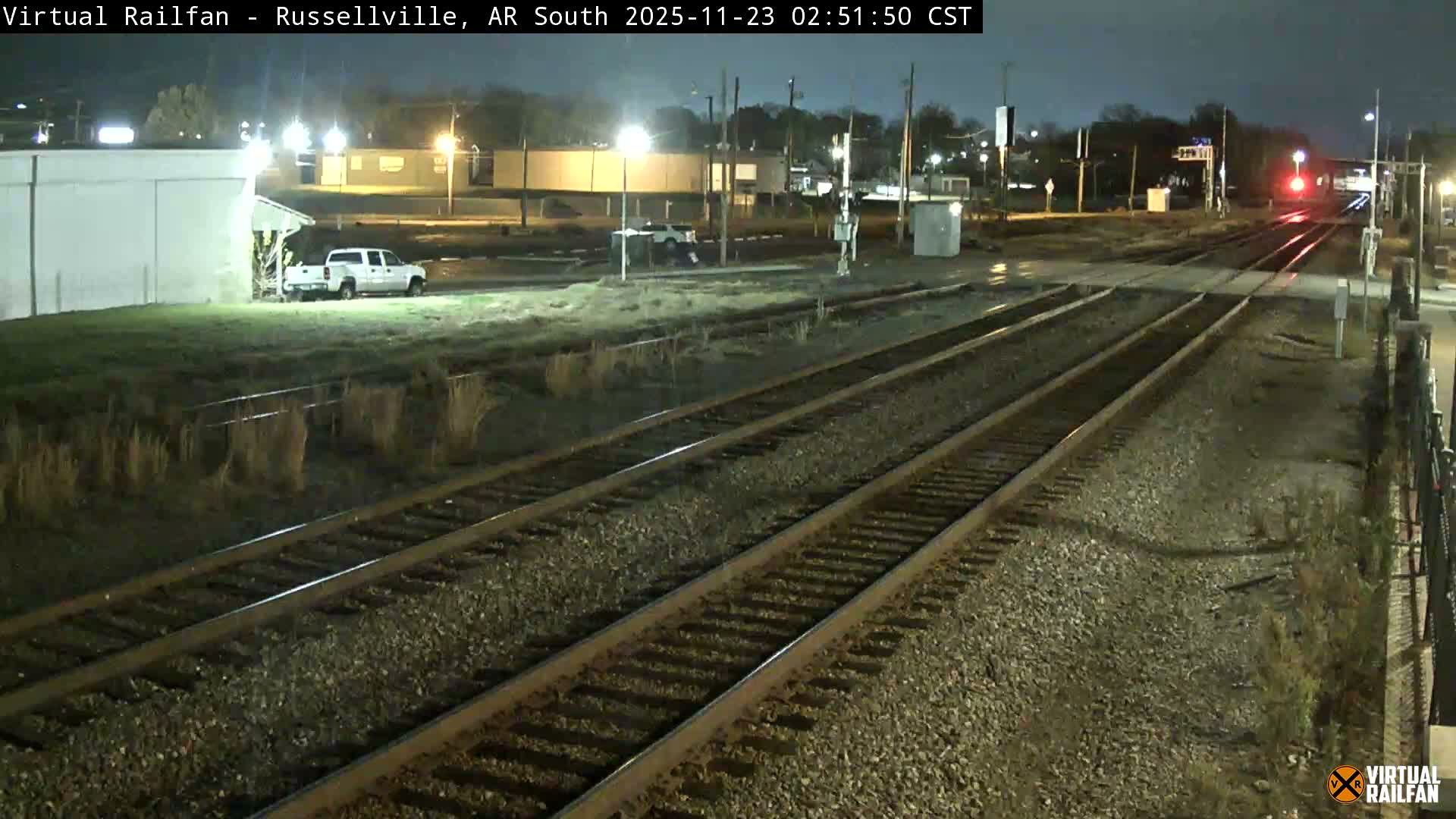 On a cloudy night, multiple train tracks recede into the distance, illuminated by streetlights that highlight a white pickup truck parked by a building and a red signal light further down the railway.