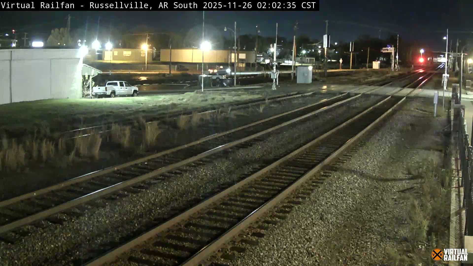 A clear night scene shows a multi-track railroad crossing with damp ground reflecting artificial lights, a white pickup truck parked near a building, and a red signal light visible in the distance on the tracks.