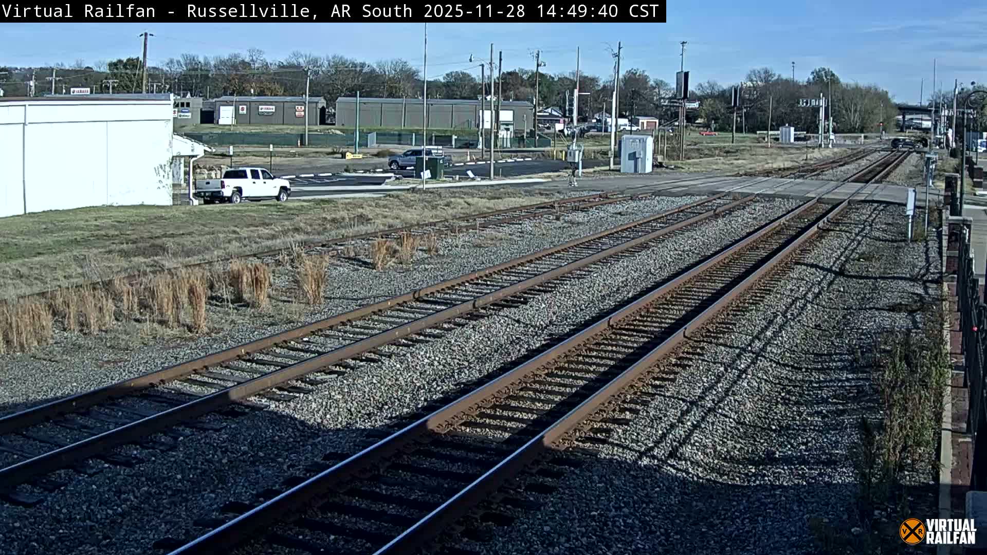 The image shows several parallel railroad tracks with gravel beds, leading towards a road crossing and distant buildings under a clear, sunny sky.