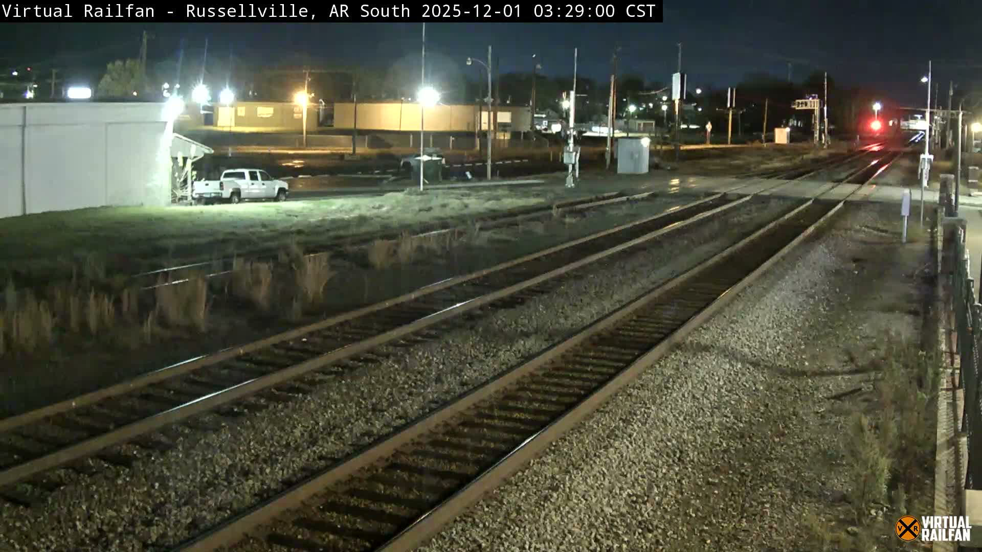 Multiple railroad tracks are illuminated by streetlights at night, with a white pickup truck parked near a building and a red signal light visible in the distance on a clear evening.