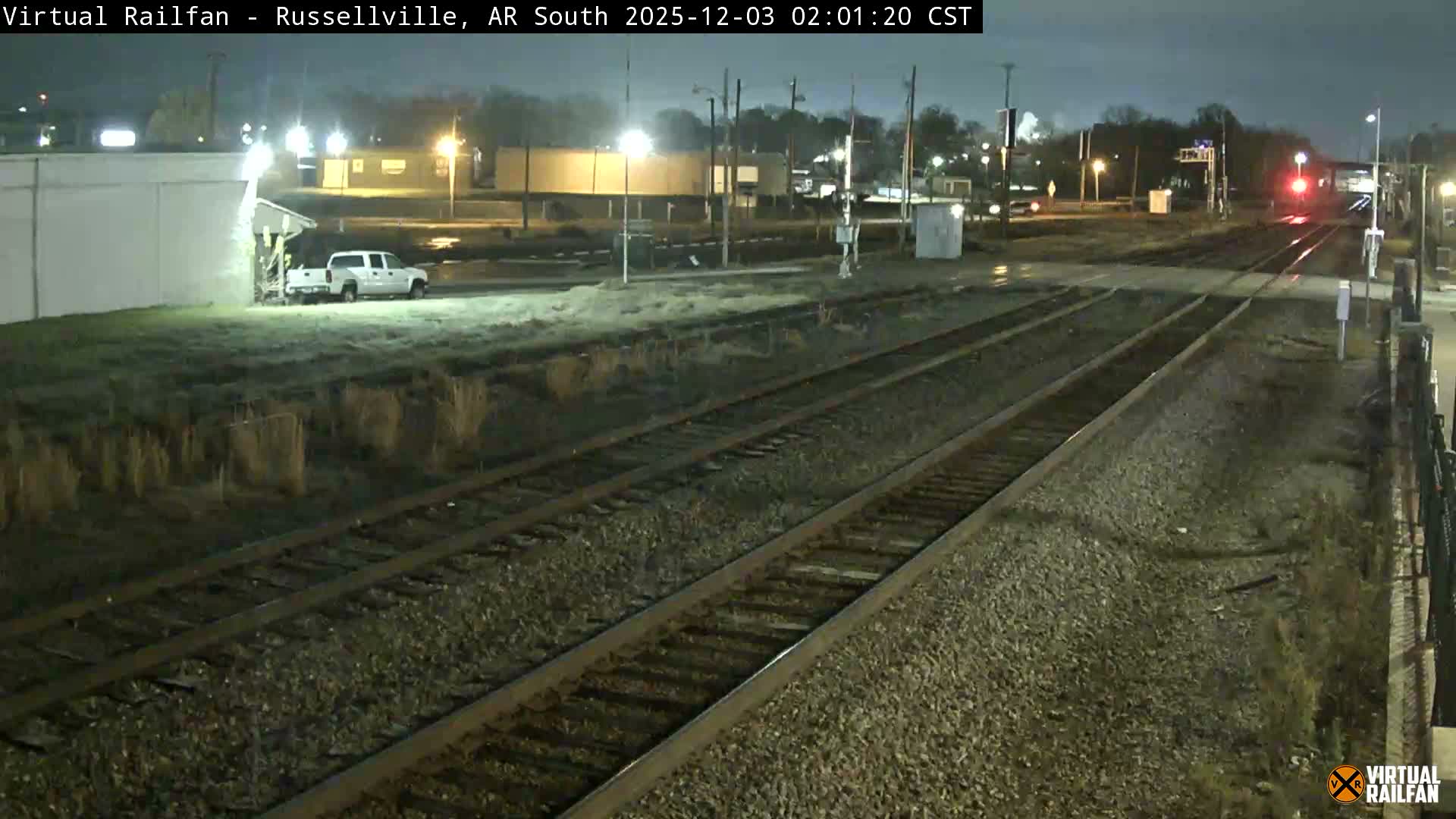 The image displays a damp, dark night scene looking down several railway tracks towards distant red signals, with a white pickup truck parked next to a building on the left.