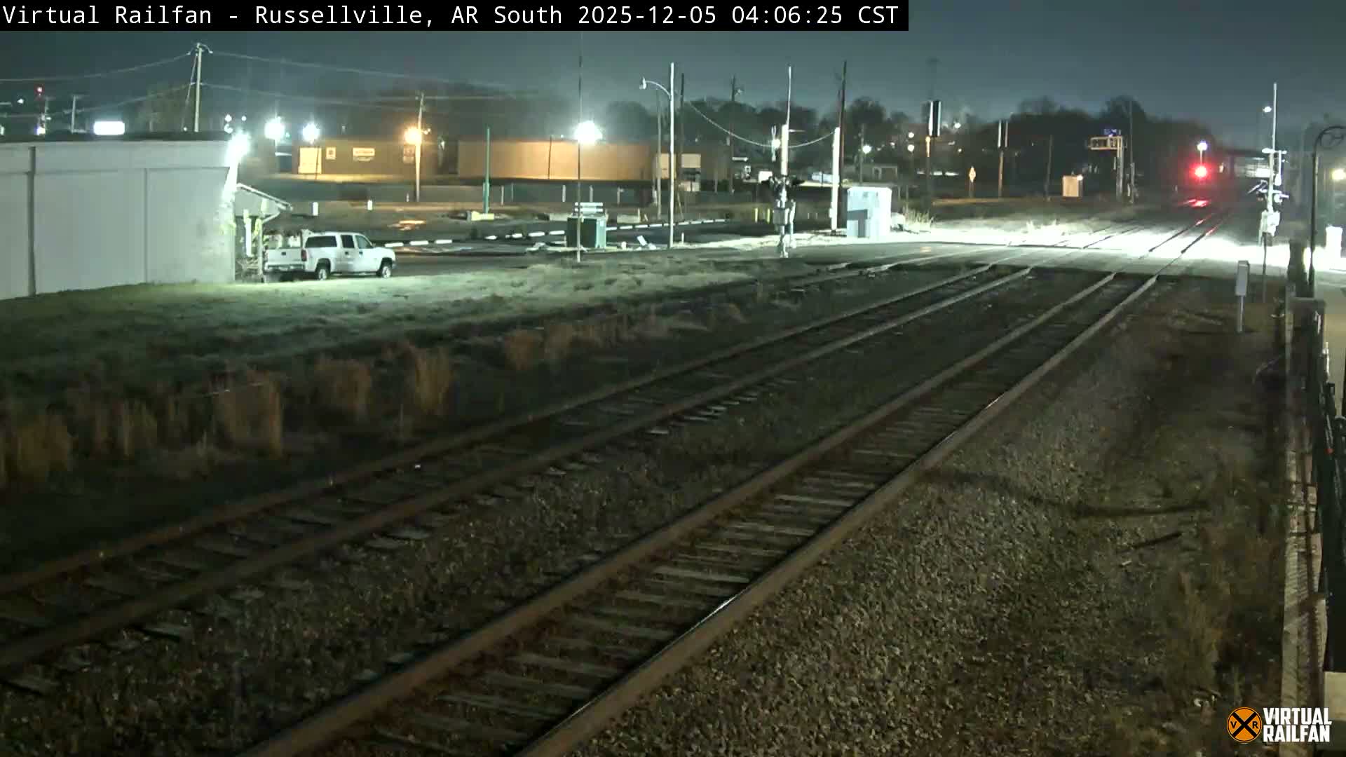 Multiple train tracks converge towards a lighted railroad crossing at night, with buildings and a white pickup truck visible in the frosty background under a clear, dark sky.