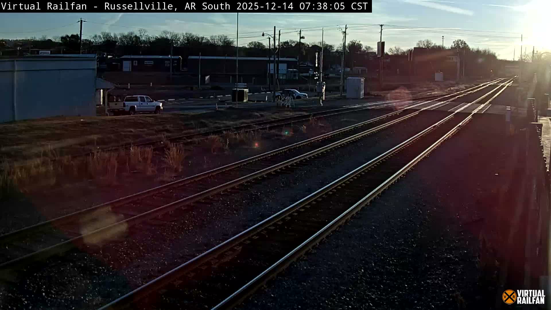 Multiple train tracks converge towards a lighted railroad crossing at night, with buildings and a white pickup truck visible in the frosty background under a clear, dark sky.