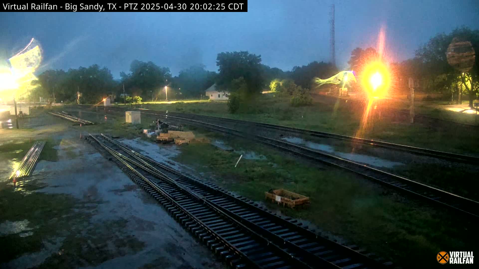 A nighttime view of rain-slicked railroad tracks and surrounding grounds, with a bright light source in the distance.