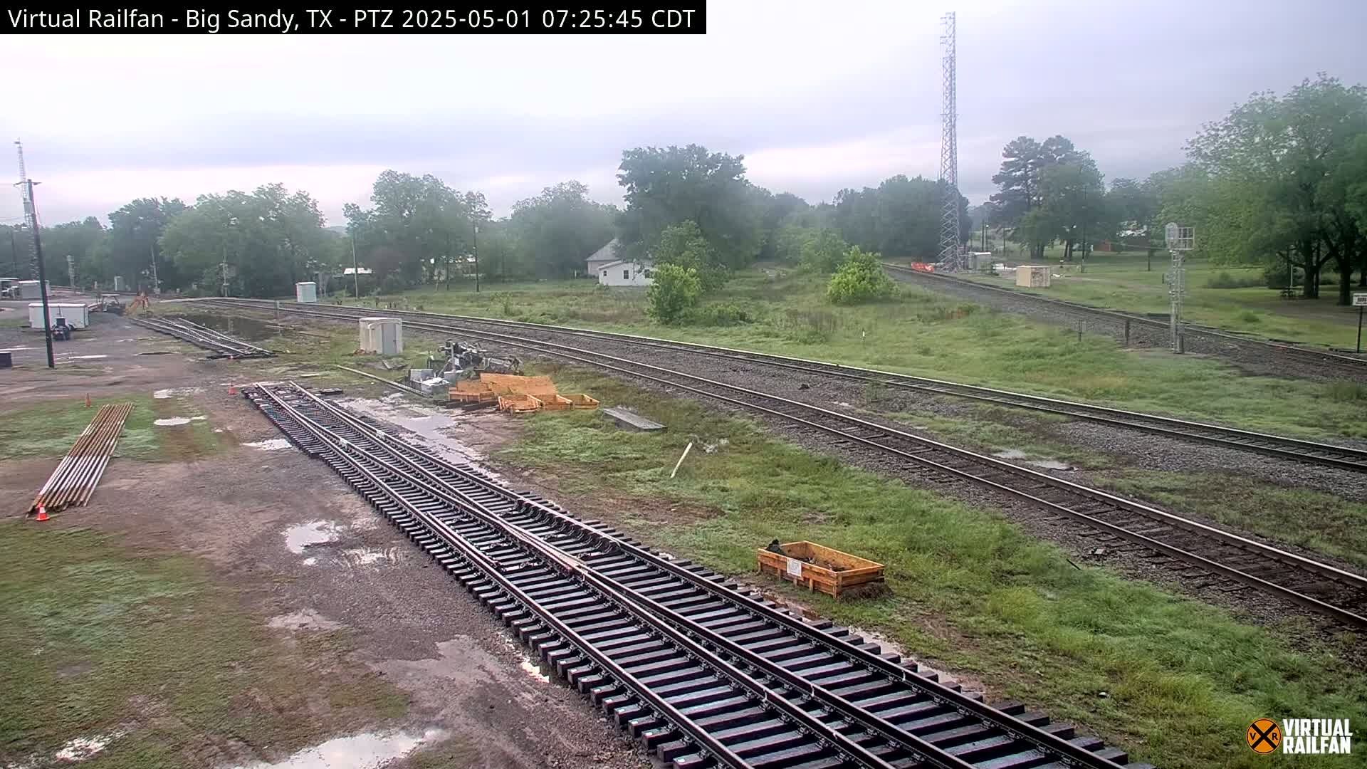 A view of railroad tracks and surrounding grassy areas on an overcast day with some puddles present.