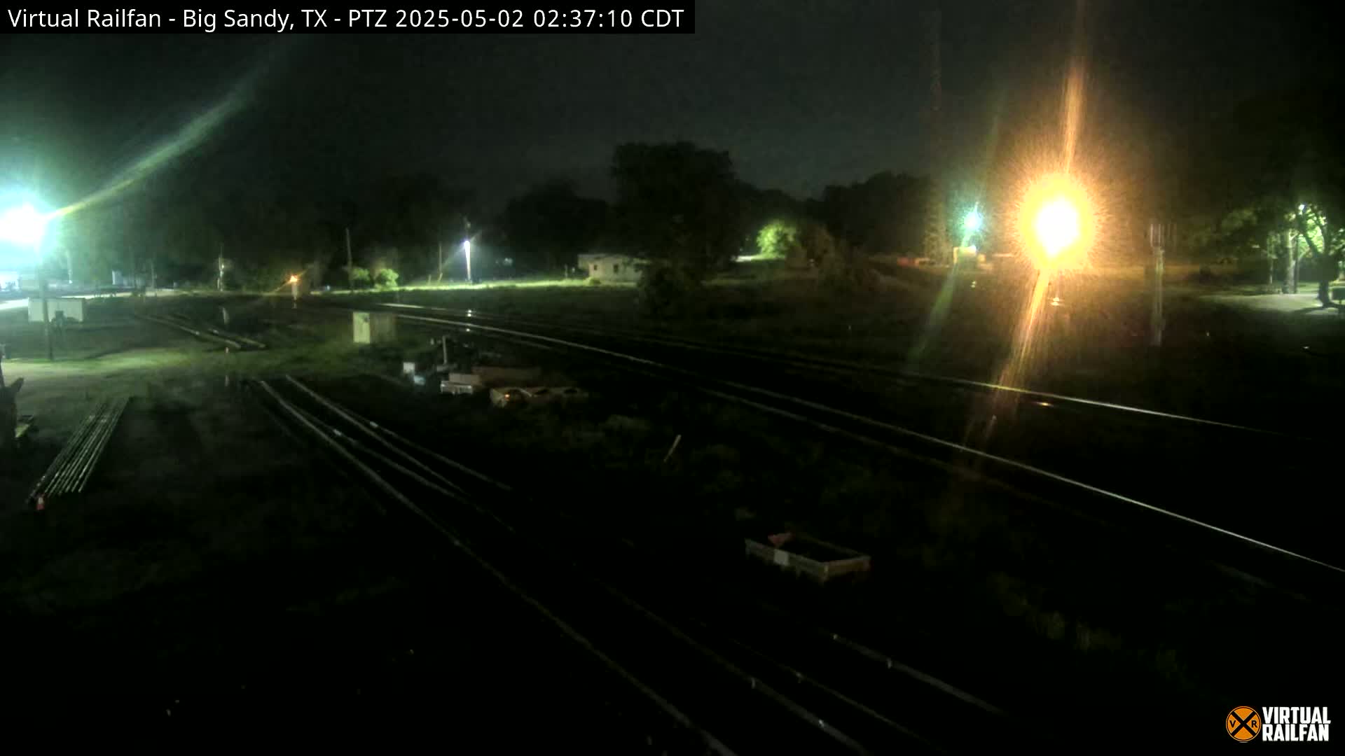 A nighttime view of railroad tracks and surrounding area, illuminated by artificial lights, with some puddles present on the ground.