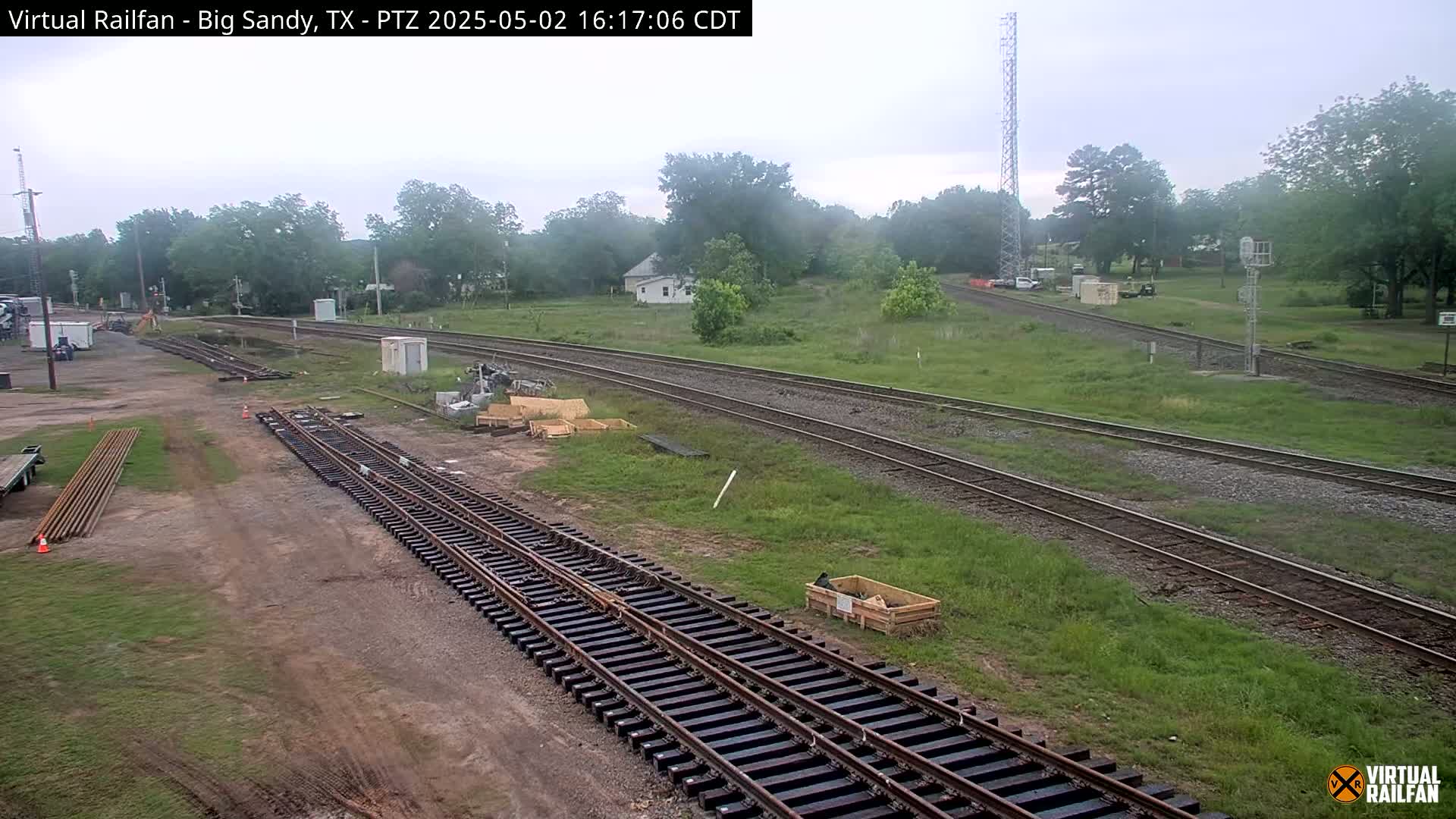 Railroad tracks under construction are visible, surrounded by grassy fields and trees under an overcast sky.
