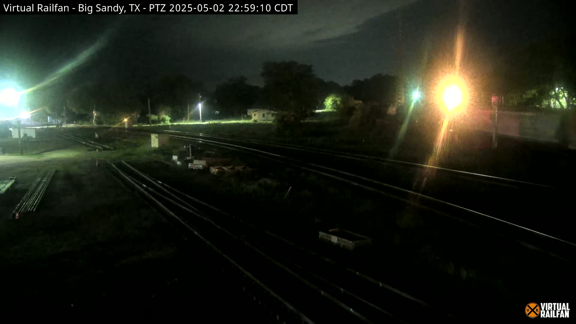 A nighttime view of railroad tracks and surrounding land, illuminated by artificial lights under a dark, possibly cloudy sky.