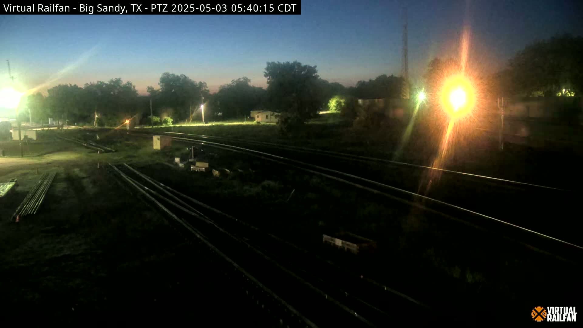 A nighttime, low-light view of several sets of railroad tracks, surrounded by dark vegetation and a few structures, under a twilight sky.