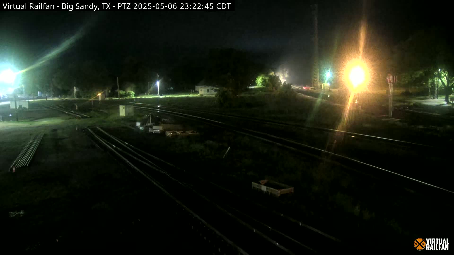 A nighttime view of railroad tracks and surrounding land, illuminated by artificial lights under hazy or foggy conditions.