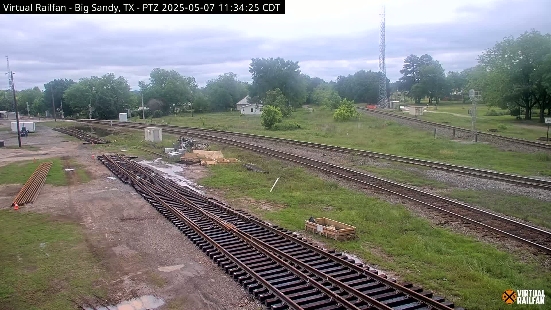 Several sets of train tracks curve through a grassy area with some construction materials and a small building visible under an overcast sky.