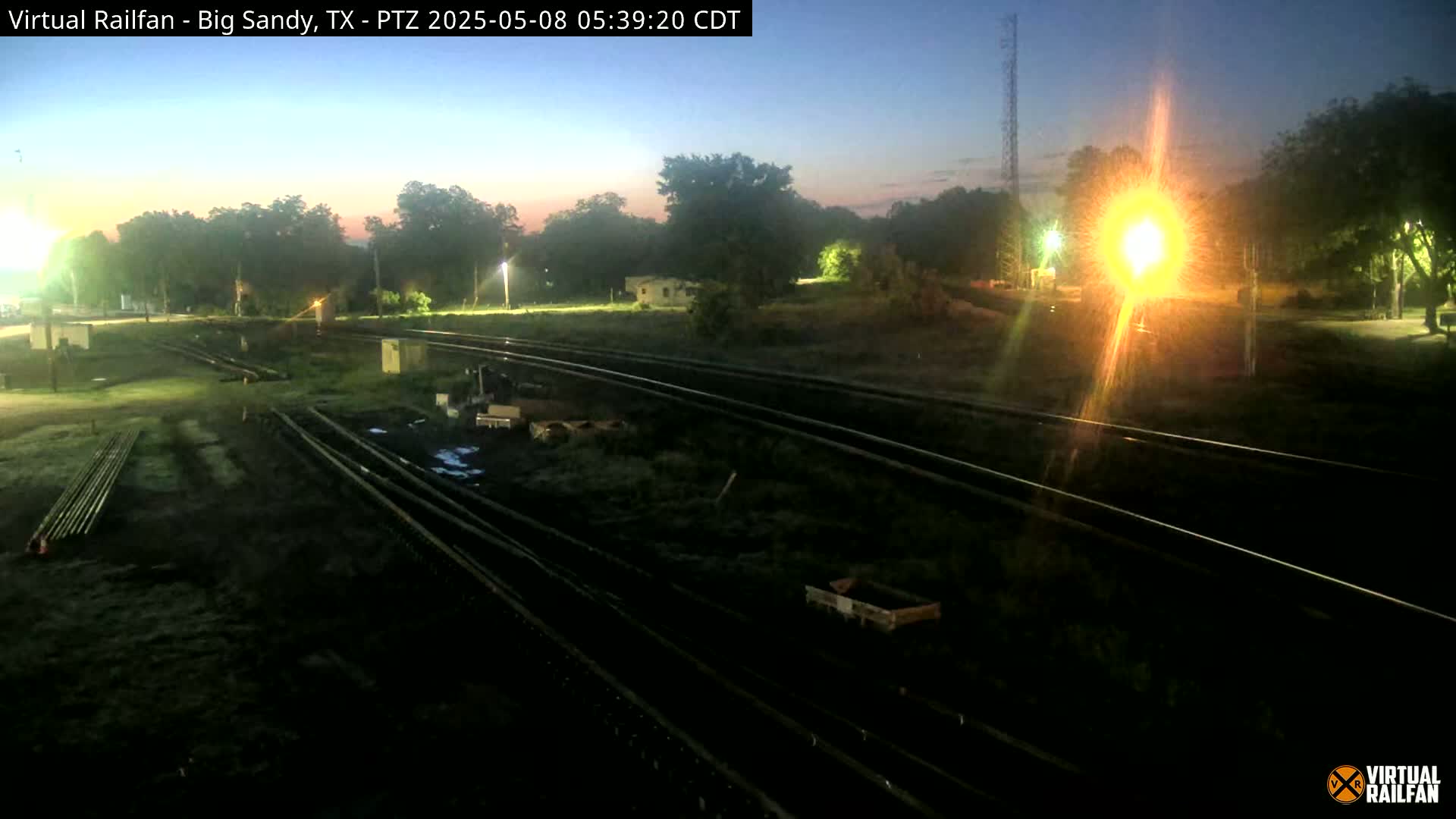 A nighttime view of a railroad yard with several sets of tracks, some equipment, and trees in the background under a twilight sky.