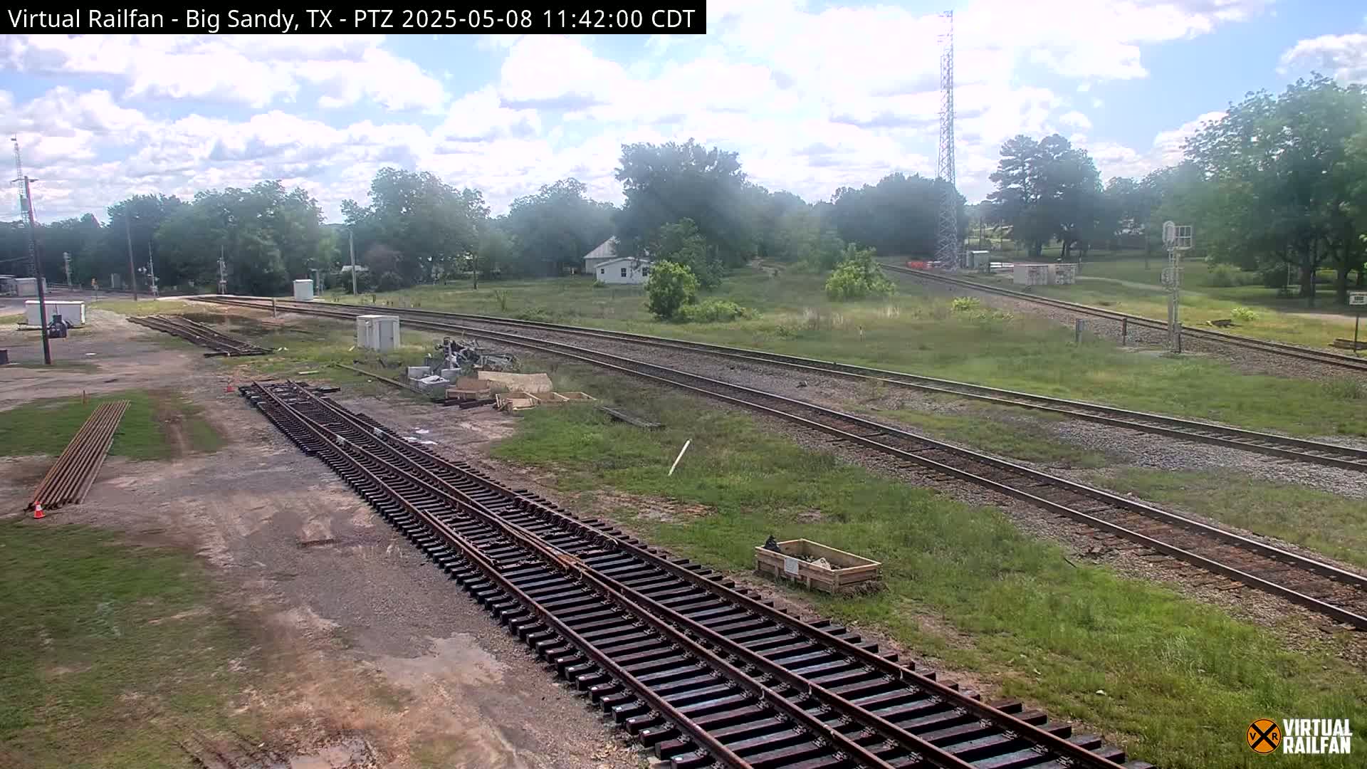 Under a partly cloudy sky, several sets of train tracks snake through a grassy area, with construction materials and equipment visible near some of the tracks.
