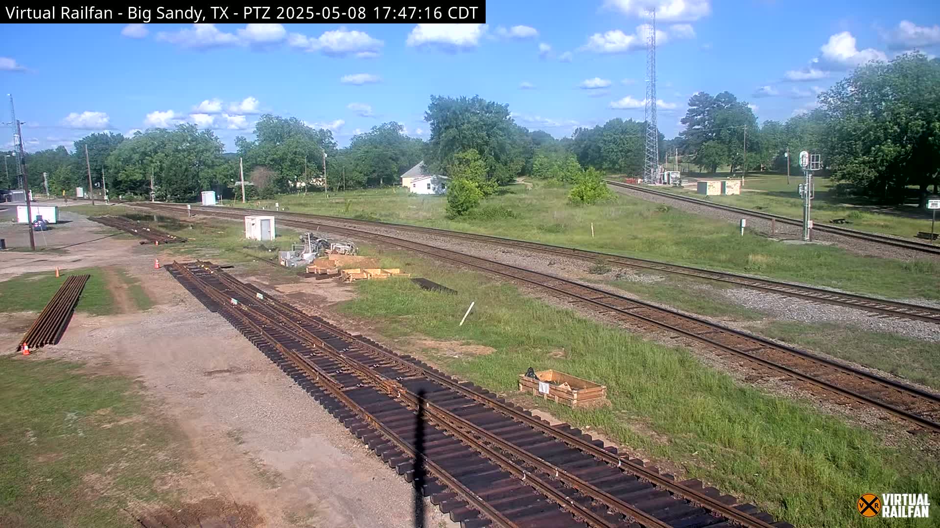 A sunny day reveals a railroad yard with multiple train tracks undergoing construction or maintenance, surrounded by grassy areas and a few buildings in the distance.