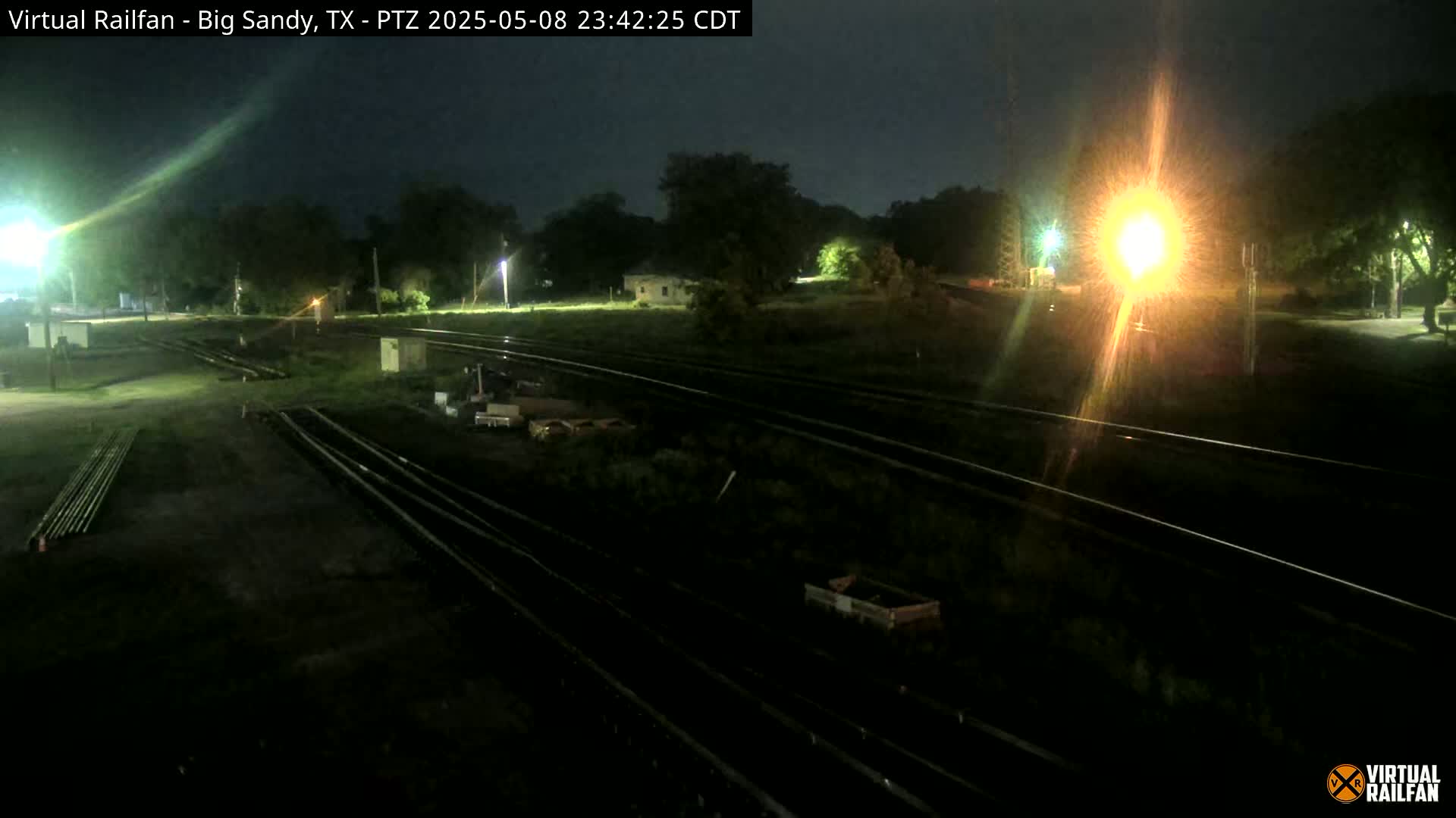 At night, under a dark sky, several sets of train tracks intersect near some small buildings and trees, with some equipment visible near the tracks.